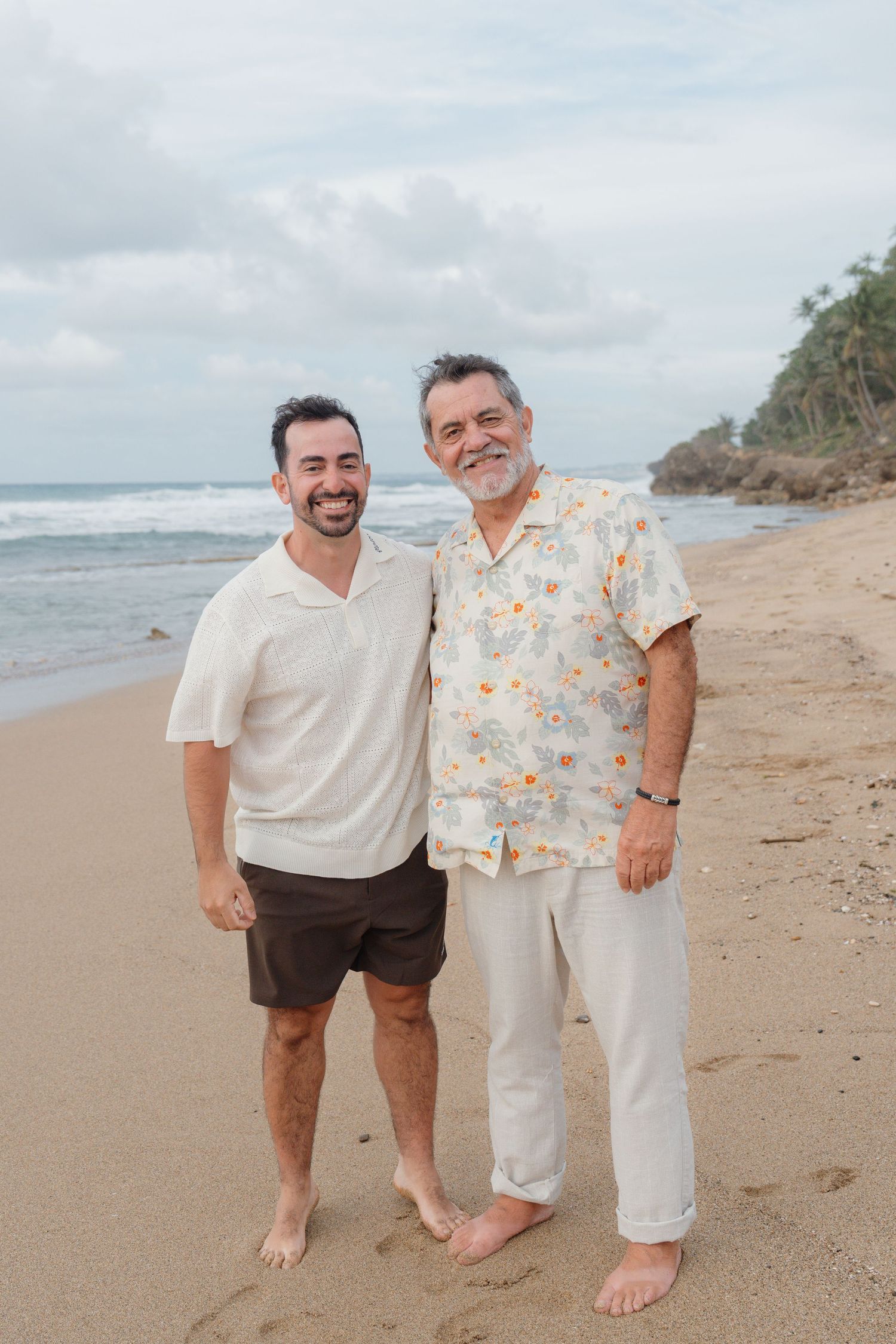 Two smiling men standing barefoot on a sandy beach with ocean waves and rocky cliffs in the background.