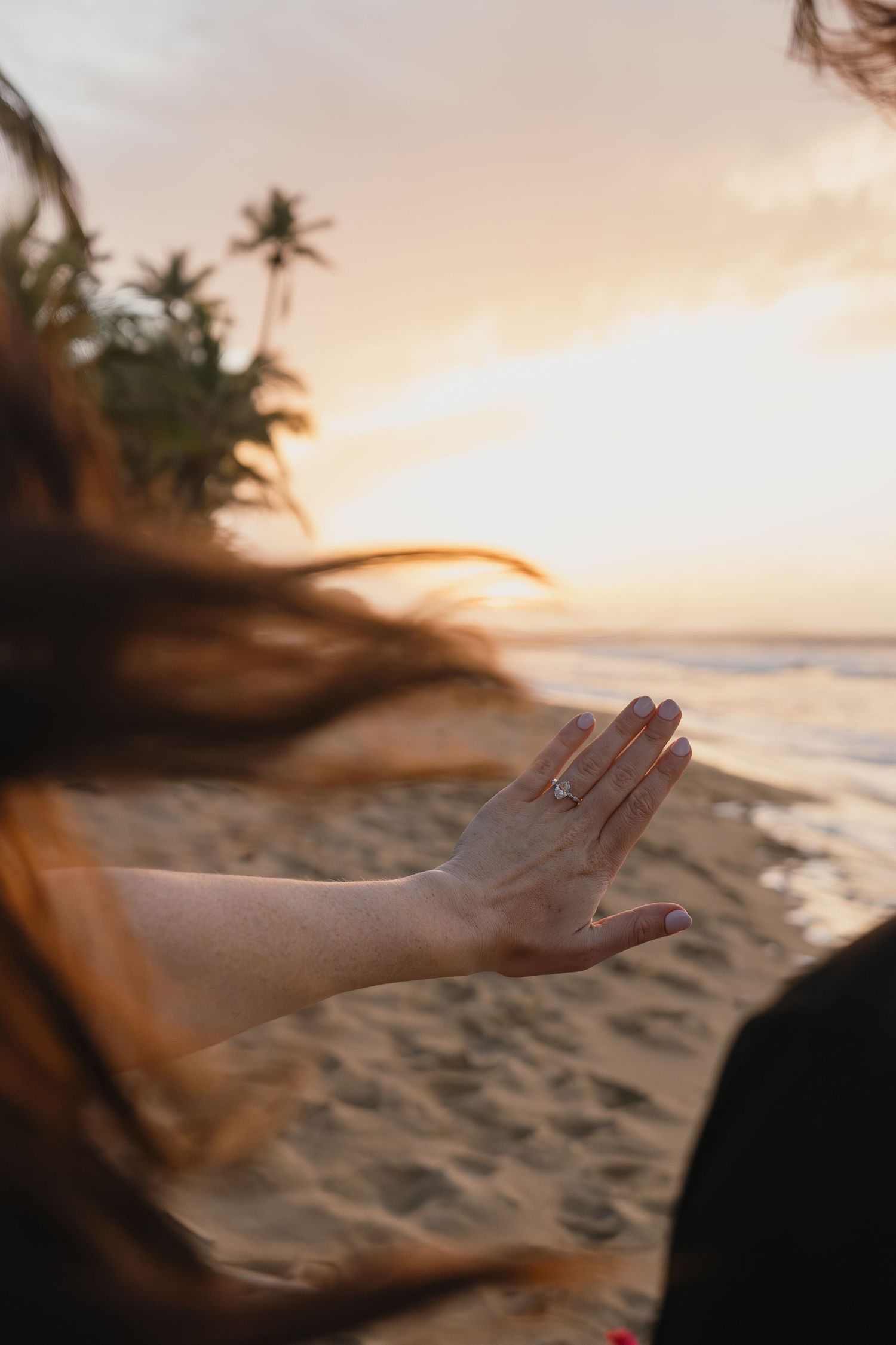 Woman's hand with ring extended on sandy beach at golden sunset with palm trees and ocean waves in background.
