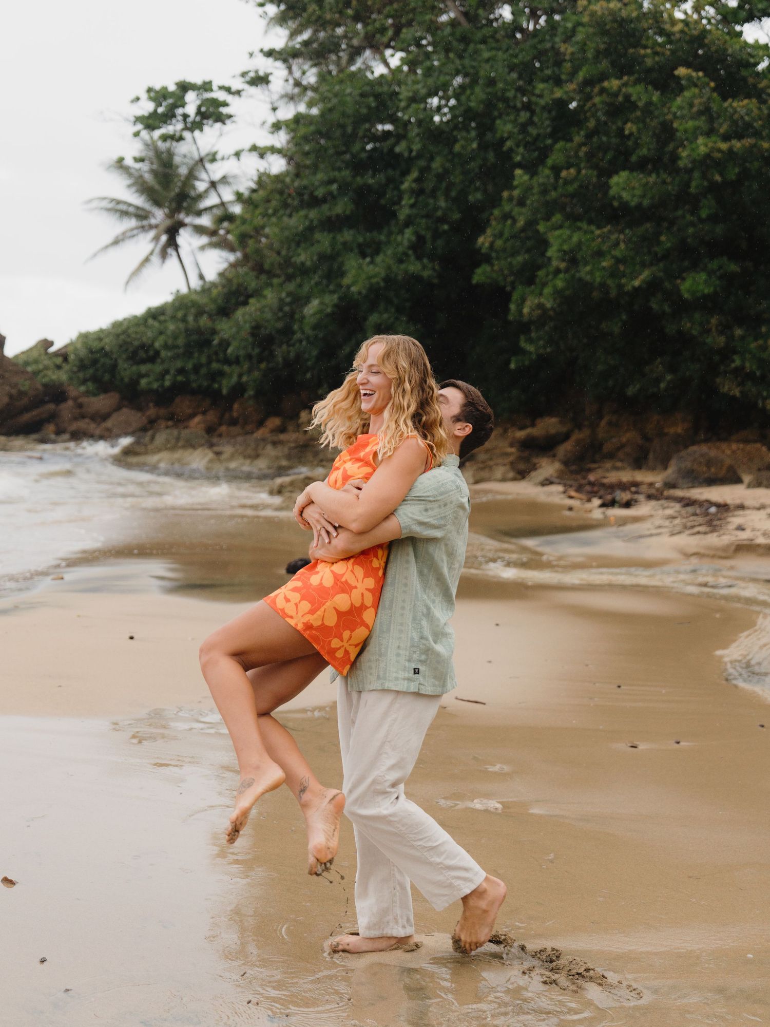 A joyful couple embraces on a tropical beach, the woman in an orange floral dress laughing as her partner lifts her.