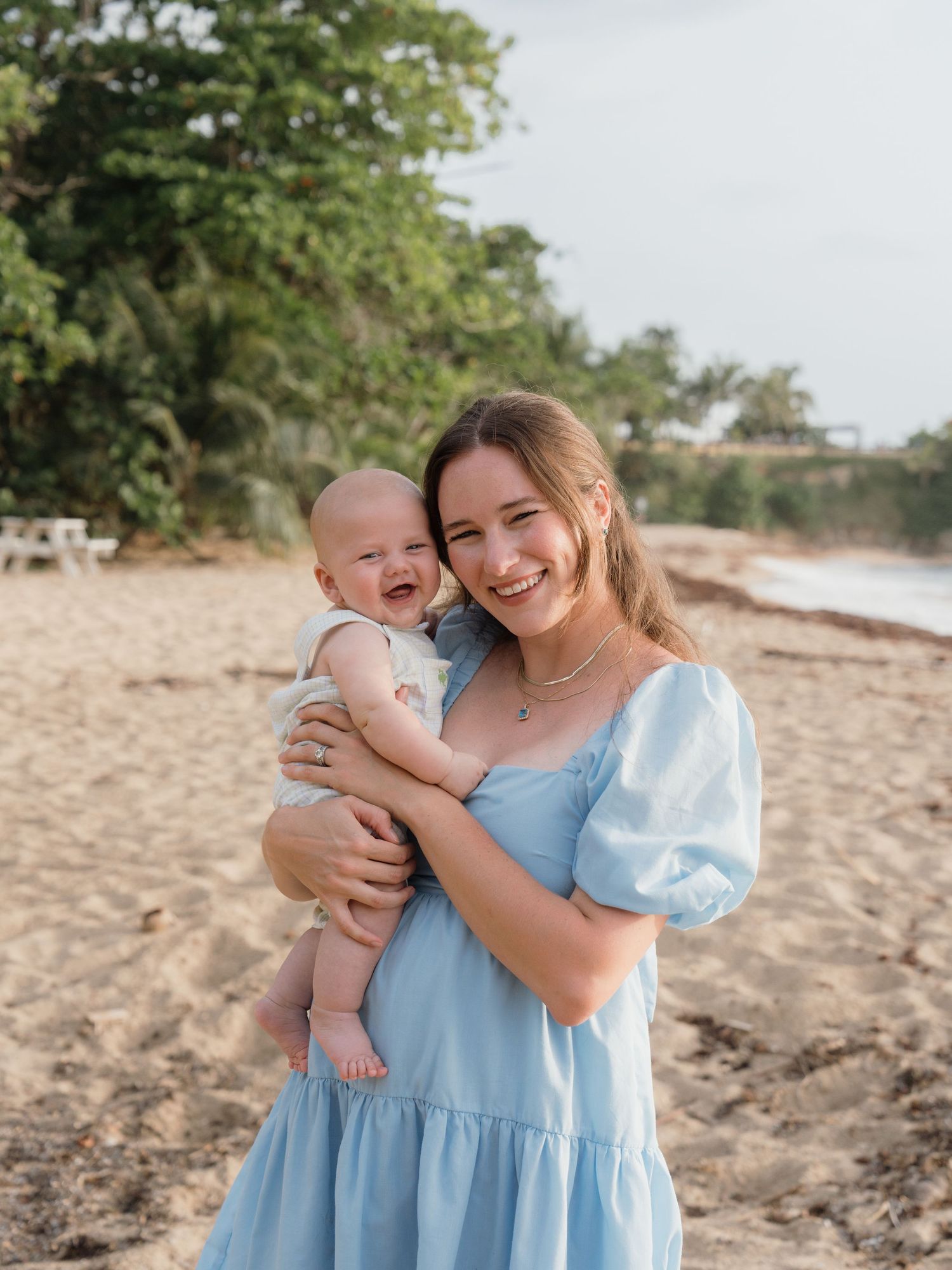 Young woman in blue dress holding a laughing baby on a sandy beach with green trees in the background.