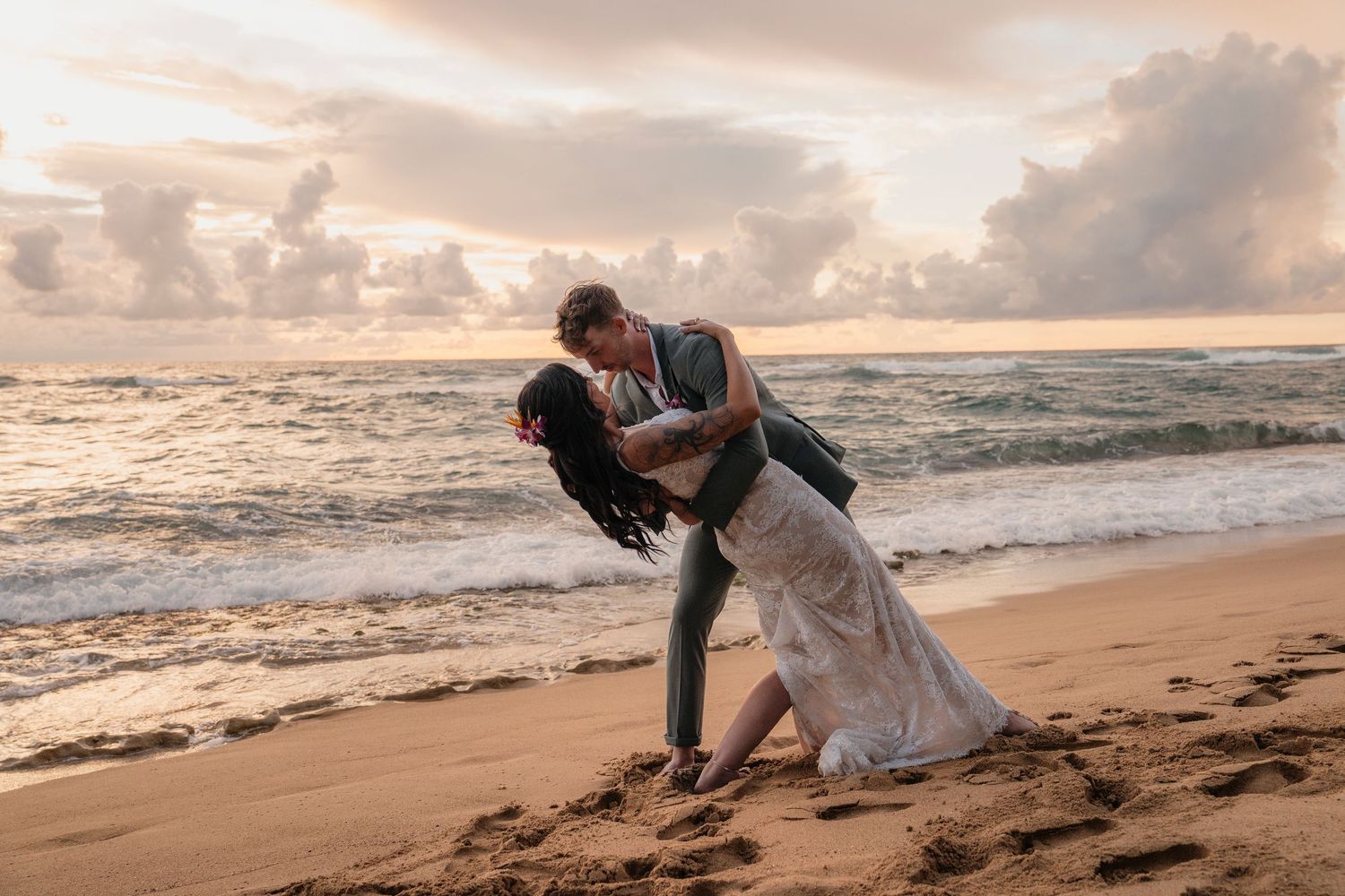Couple sharing a romantic dip kiss on a sandy beach at sunset, bride in white gown, groom in gray suit.