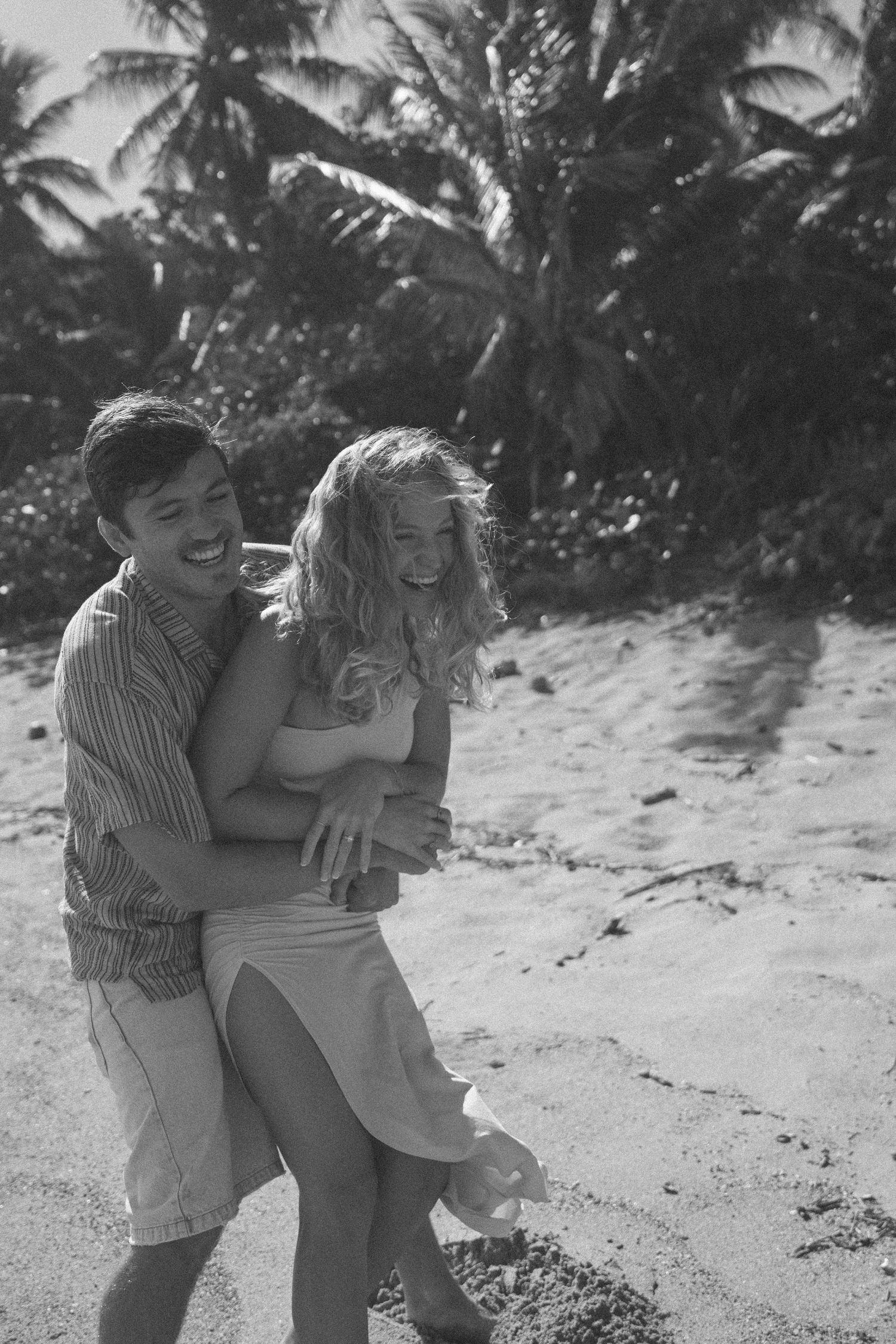 Happy couple embracing and laughing on a tropical sandy beach surrounded by palm trees in black and white.