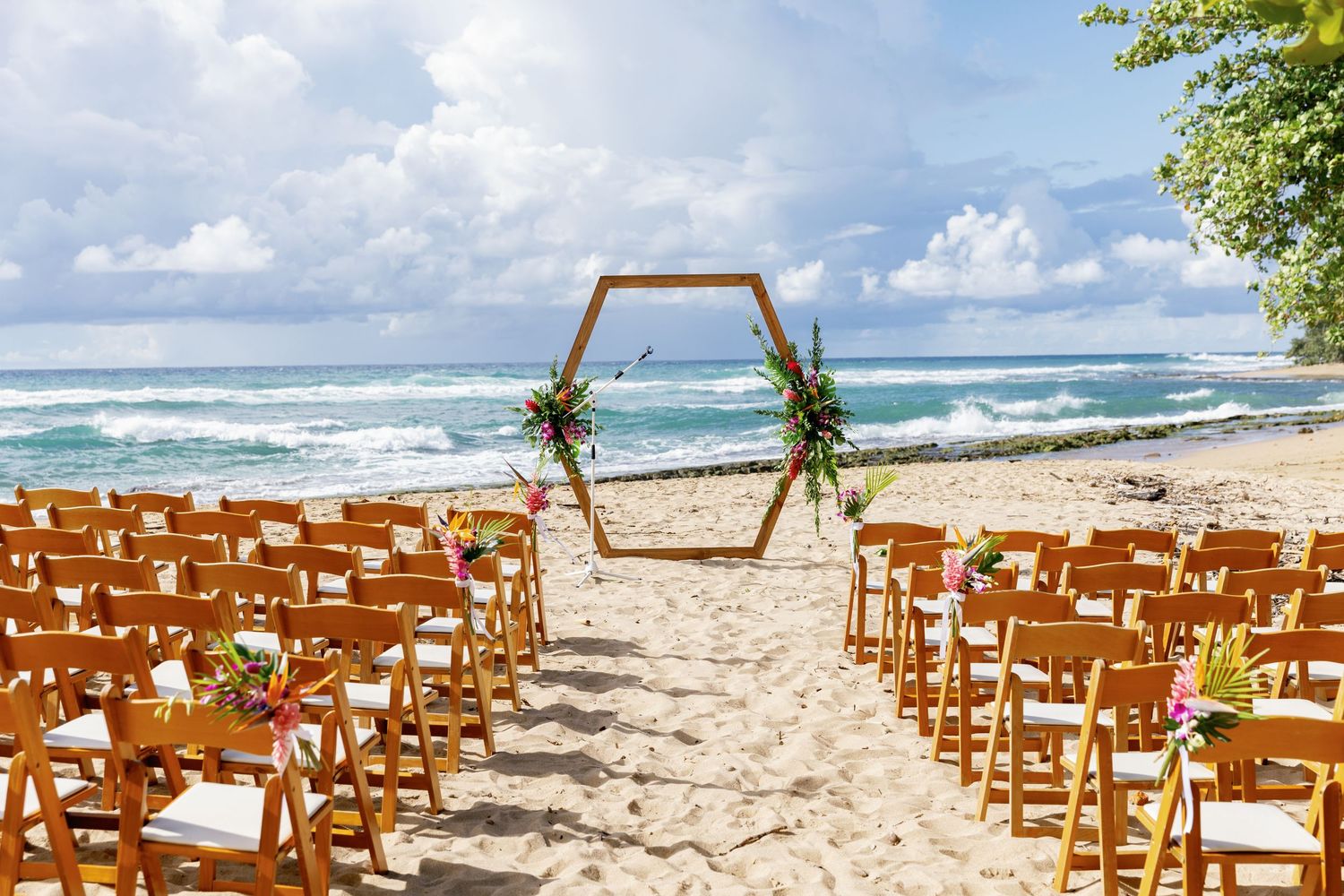 Elegant beach wedding setup with wooden chairs and hexagonal floral arch overlooking ocean waves.