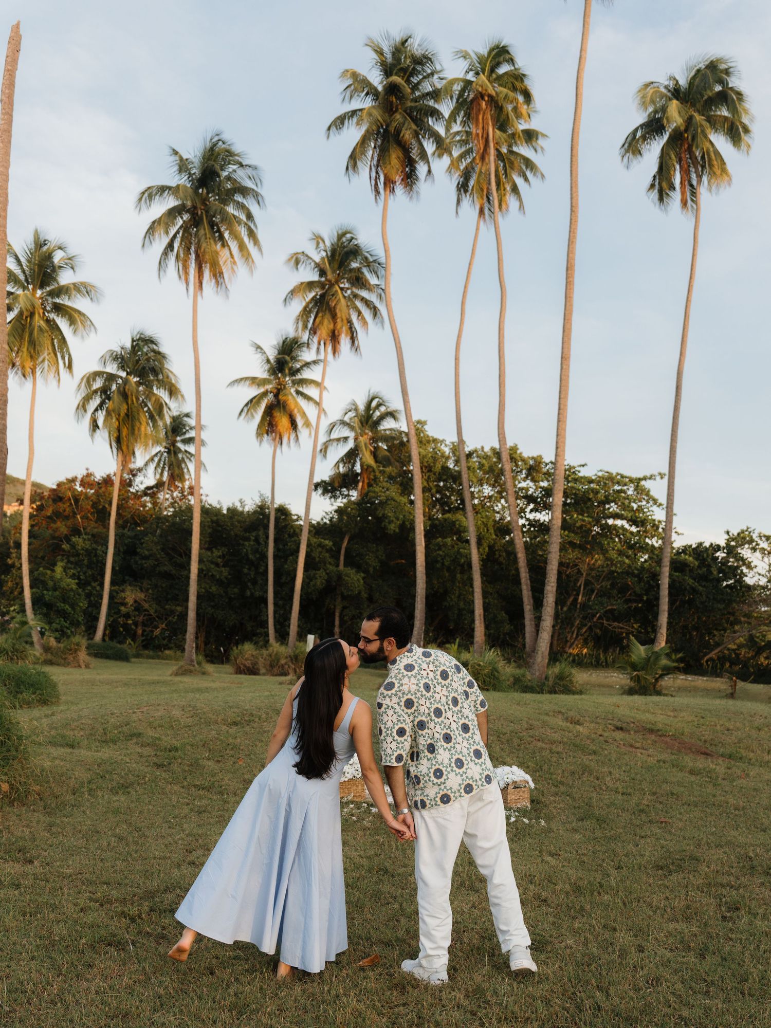 A couple holds hands among tall palm trees on a lush green lawn at golden hour in a tropical setting.