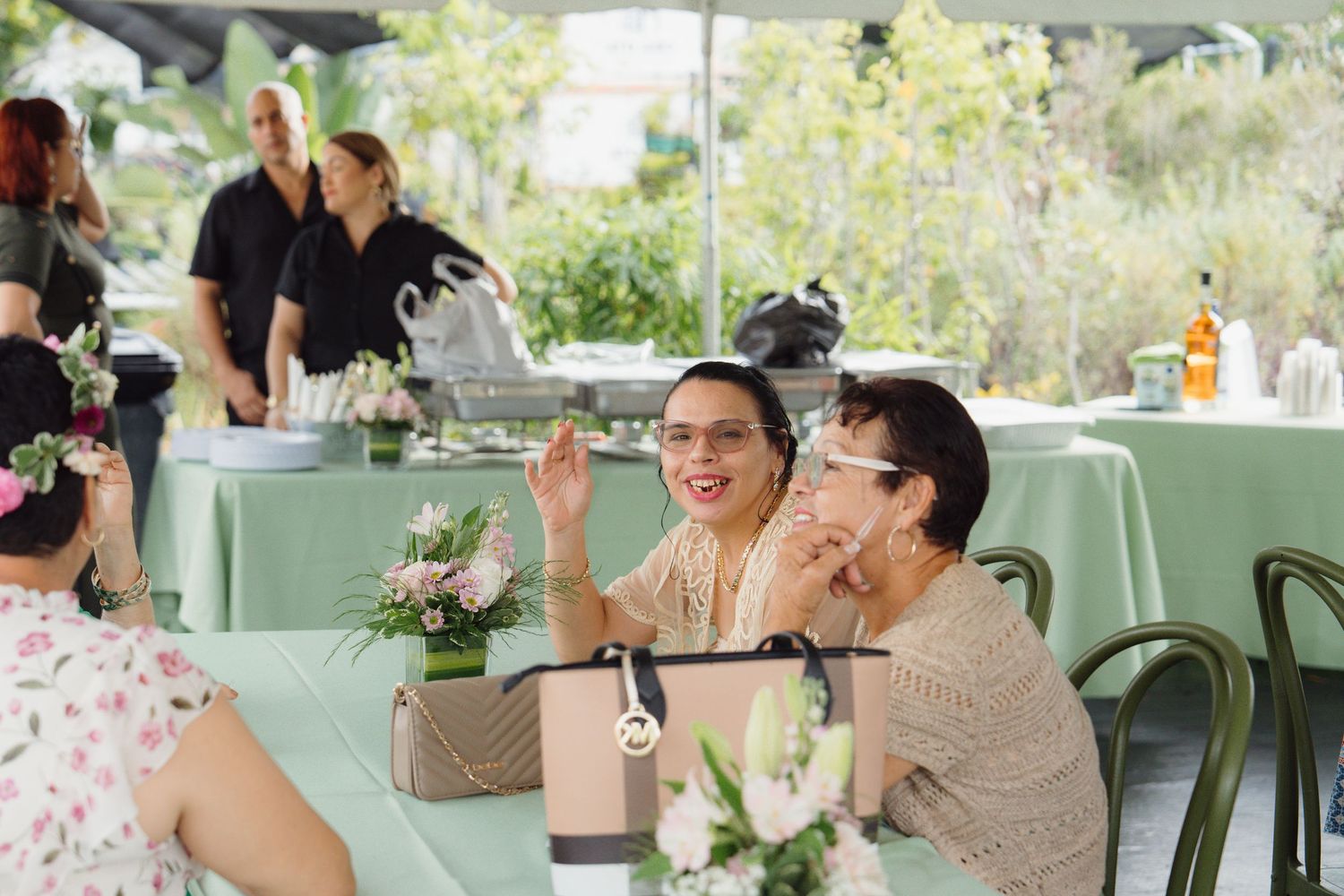 Women laughing and socializing at an outdoor event with green tablecloths and floral centerpieces.