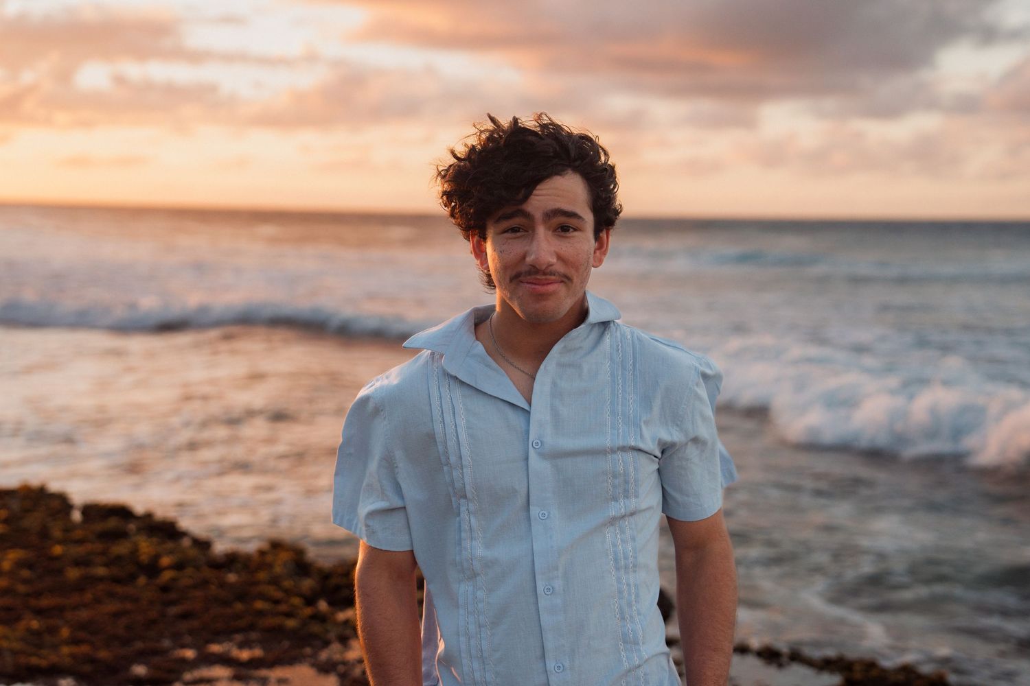Young man in light blue shirt smiling at rocky beach during golden sunset with ocean waves behind him.