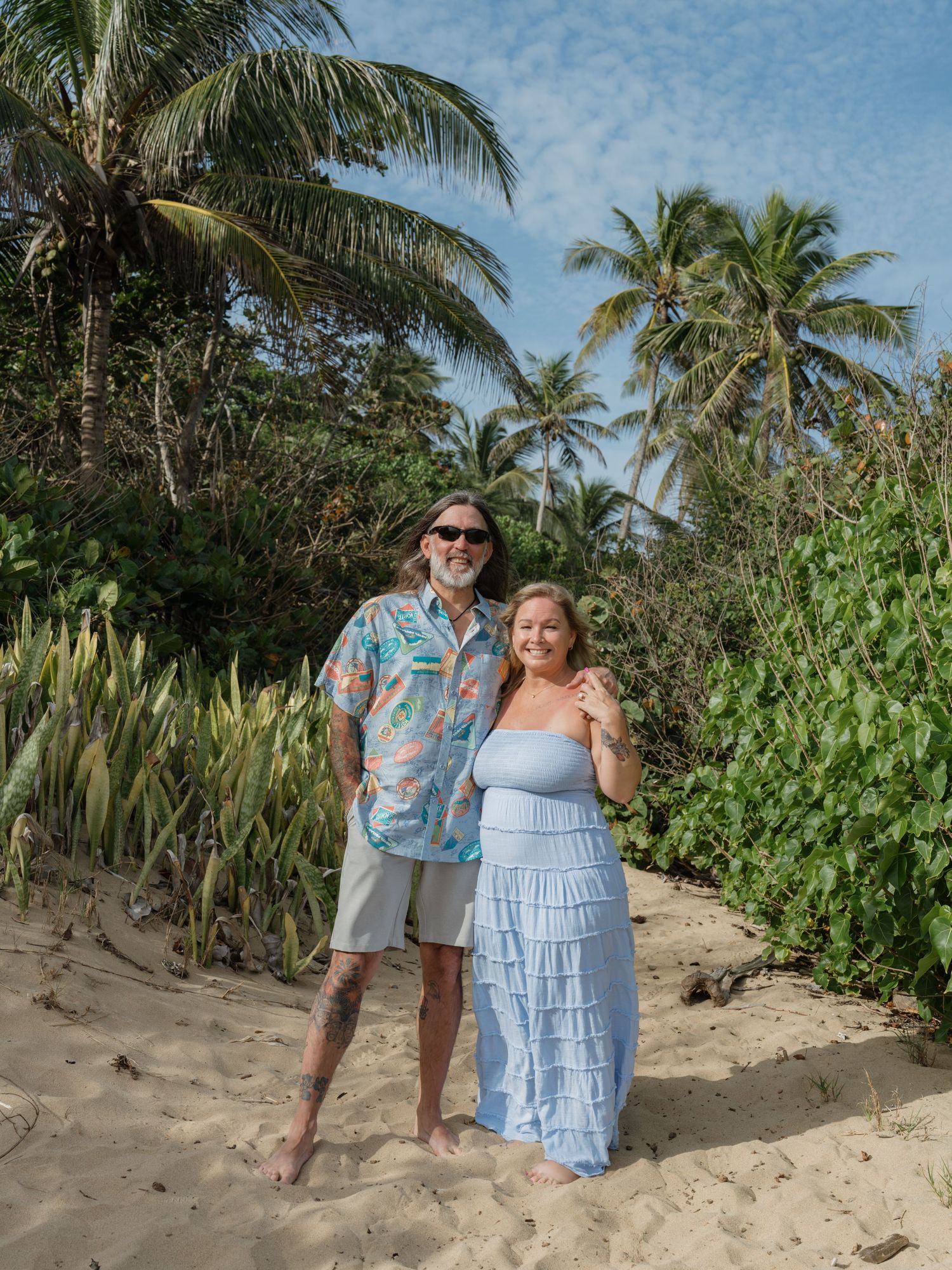 Couple posing together on sandy beach surrounded by lush tropical palm trees on a sunny day.