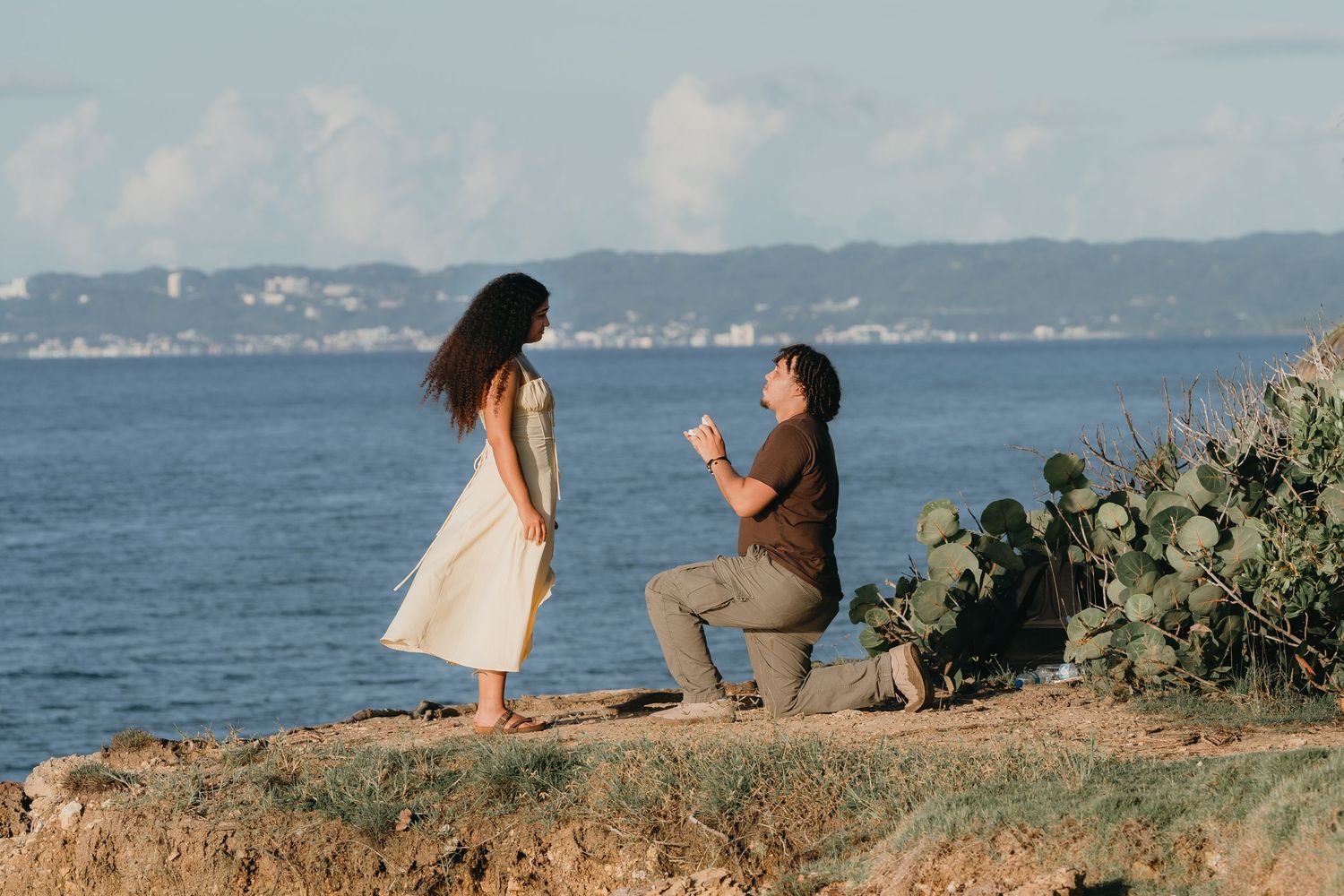 Man proposing on one knee to woman on rocky coastal cliff with ocean and blue sky in background.