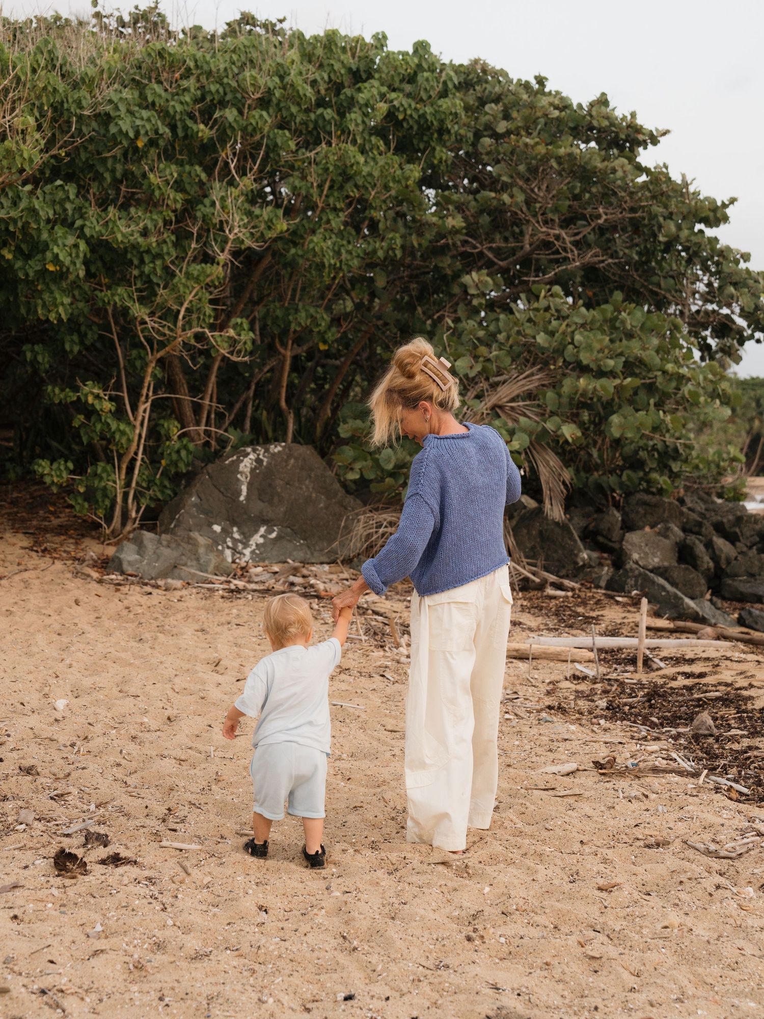 A woman and toddler walk hand-in-hand on a sandy beach toward lush green trees and rocks.