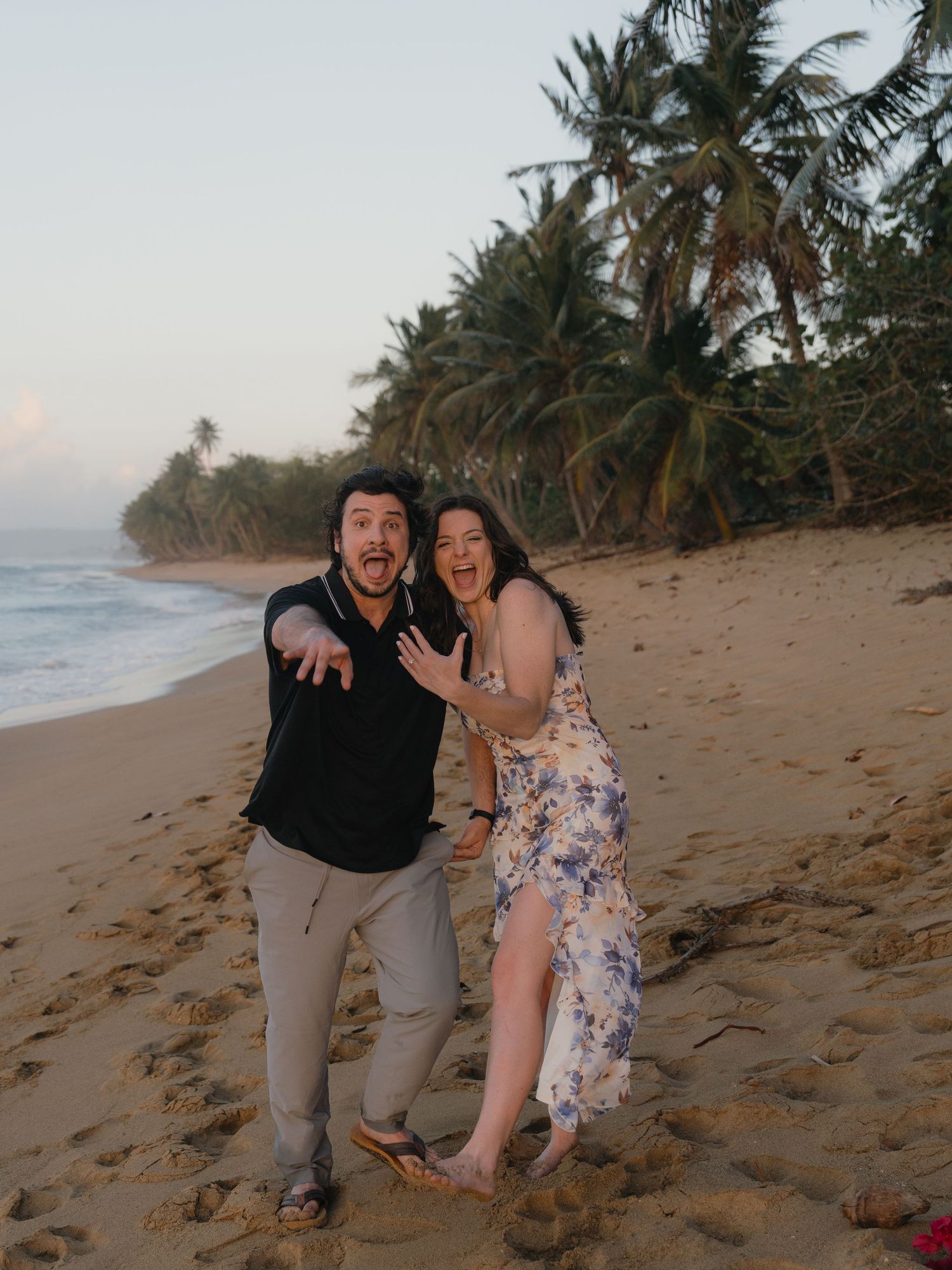 Excited couple posing on a tropical beach at sunset with palm trees and ocean waves in the background.