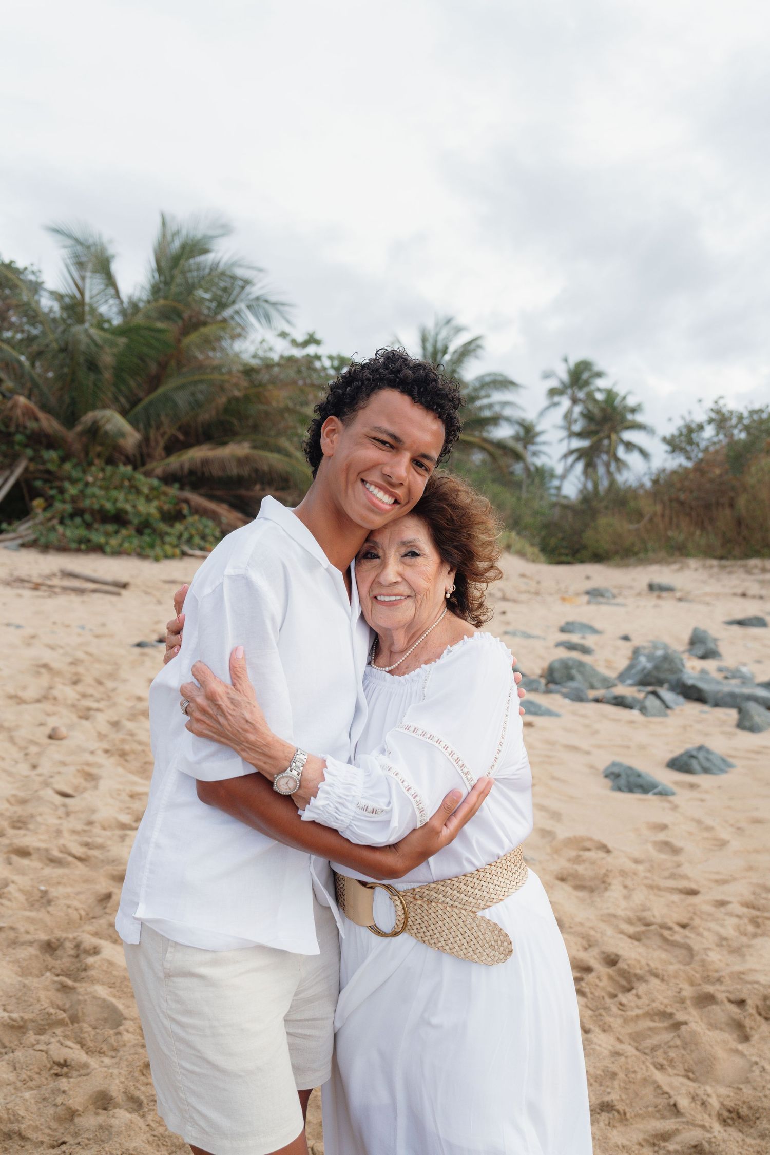Two people in white outfits embrace on a sandy beach with palm trees, smiling warmly at the camera.