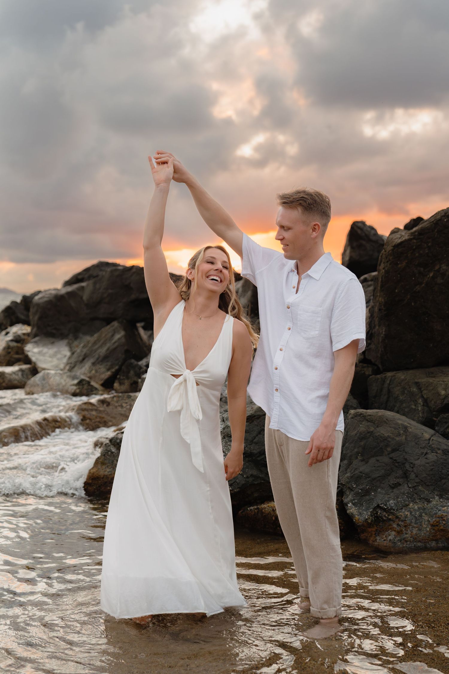 Couple dancing on rocky beach at sunset, woman in white dress laughing as man spins her hand.