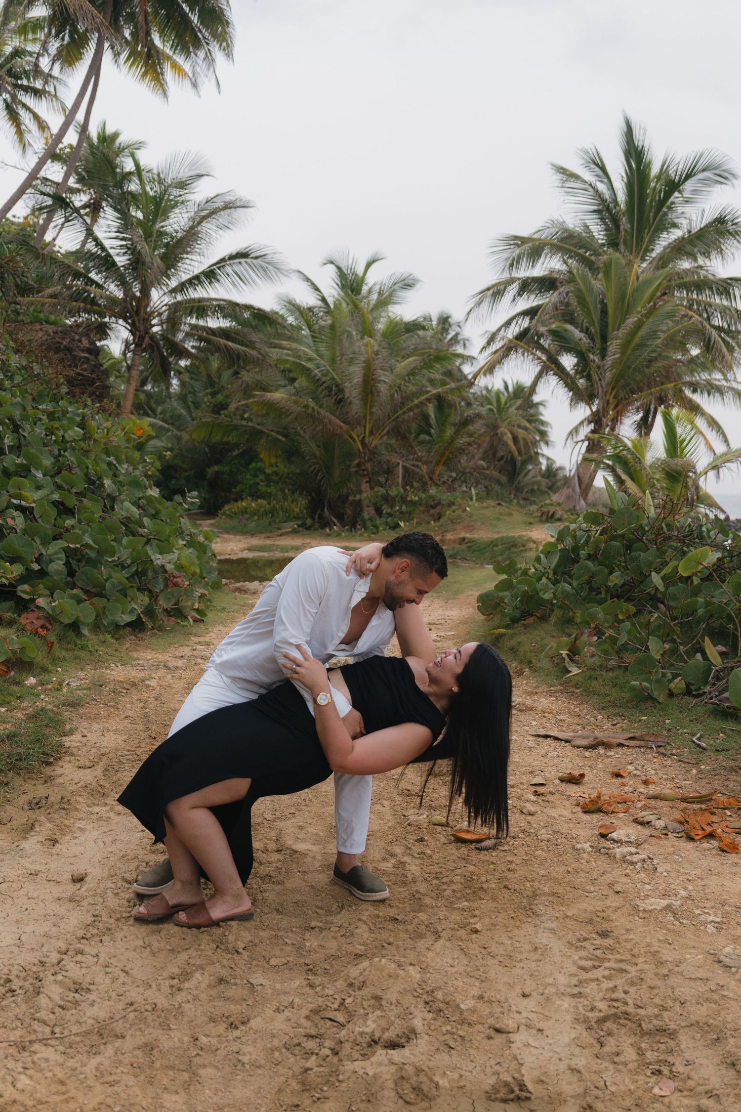 Couple sharing a romantic dip kiss on a sandy tropical path surrounded by lush palm trees.