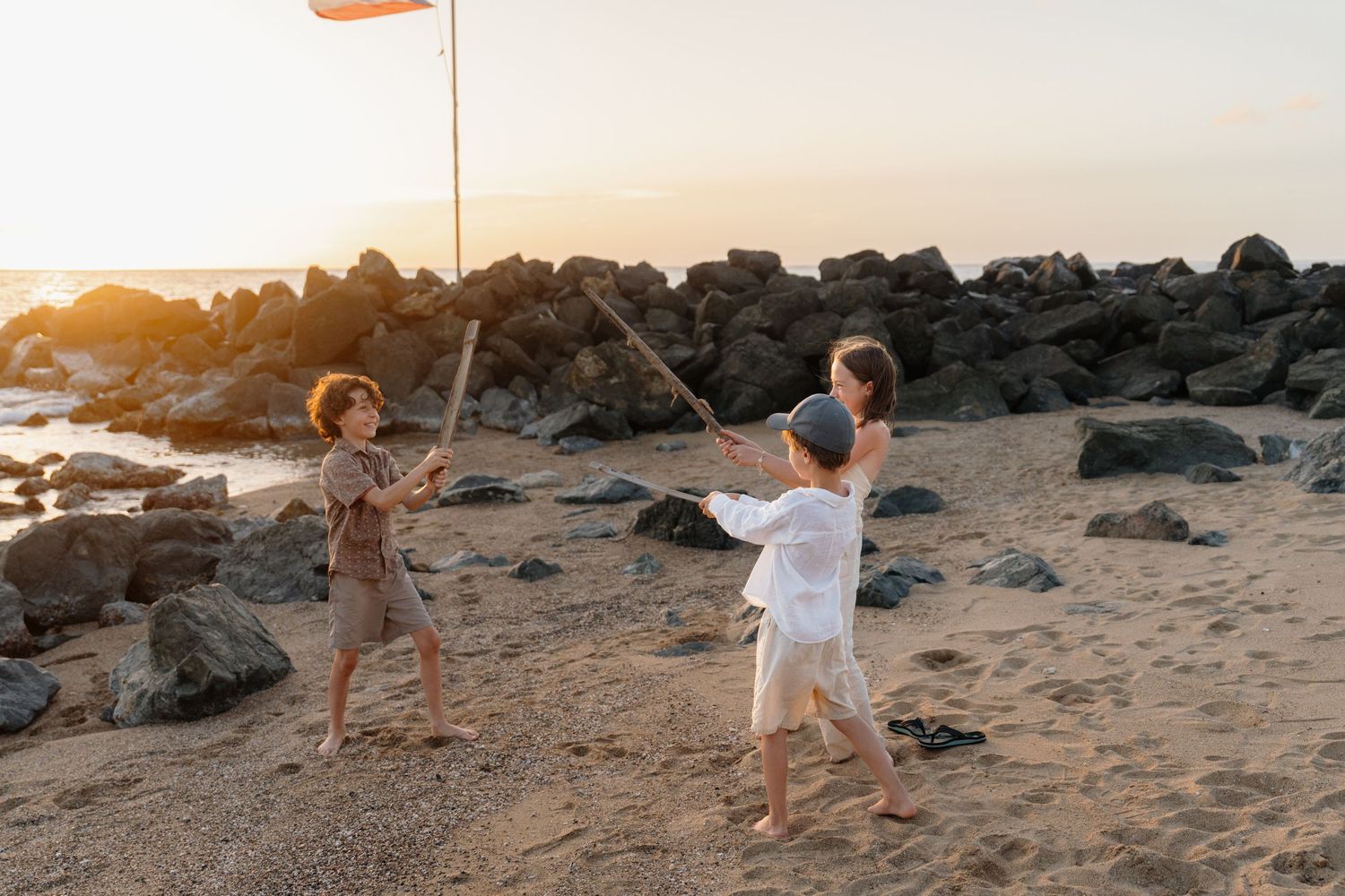 Three children playing with sticks and a flag on a sandy beach at sunset with rocky shoreline in background.