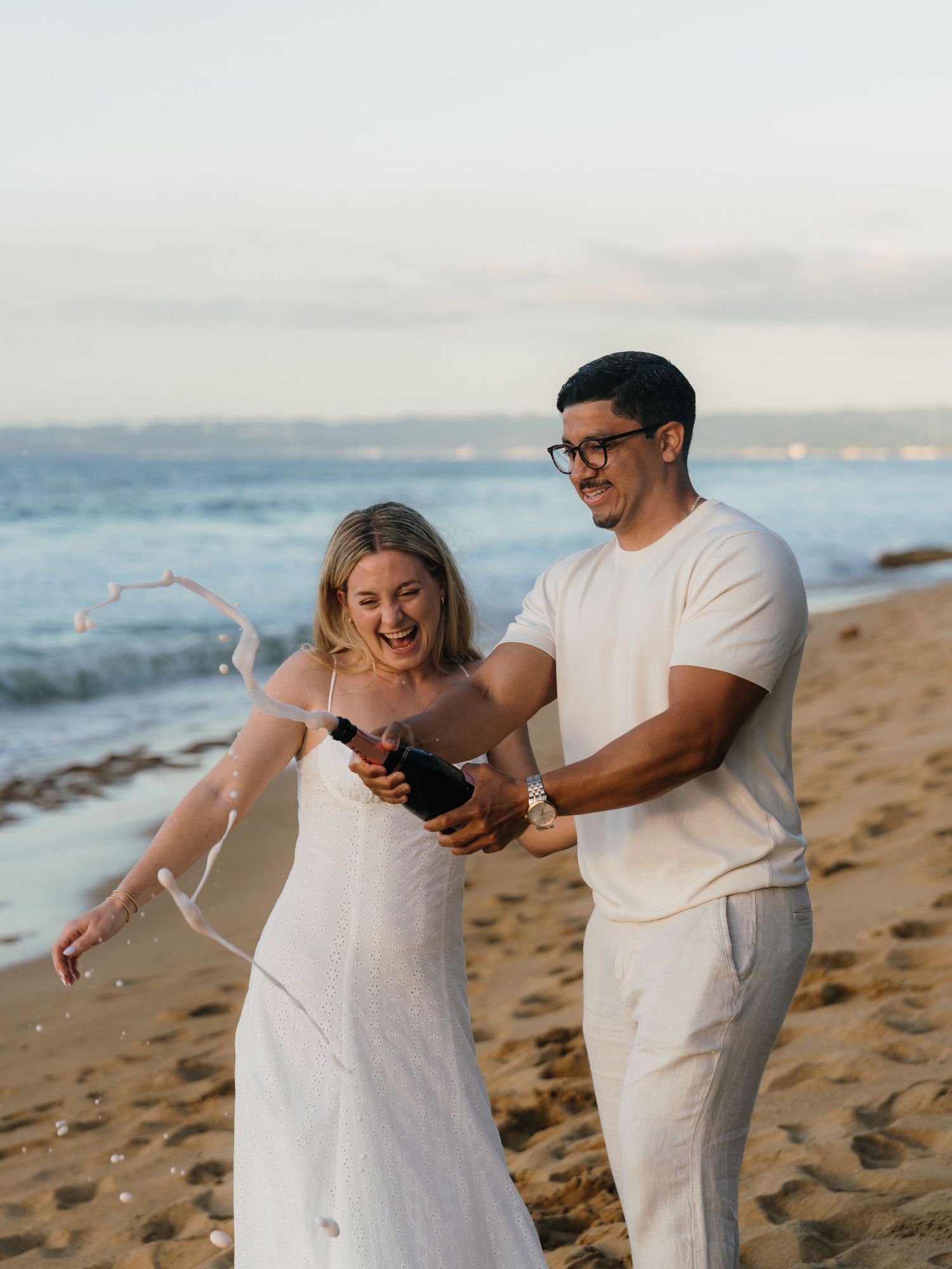 Couple celebrating on a beach by popping champagne, wearing white outfits with ocean waves in the background.