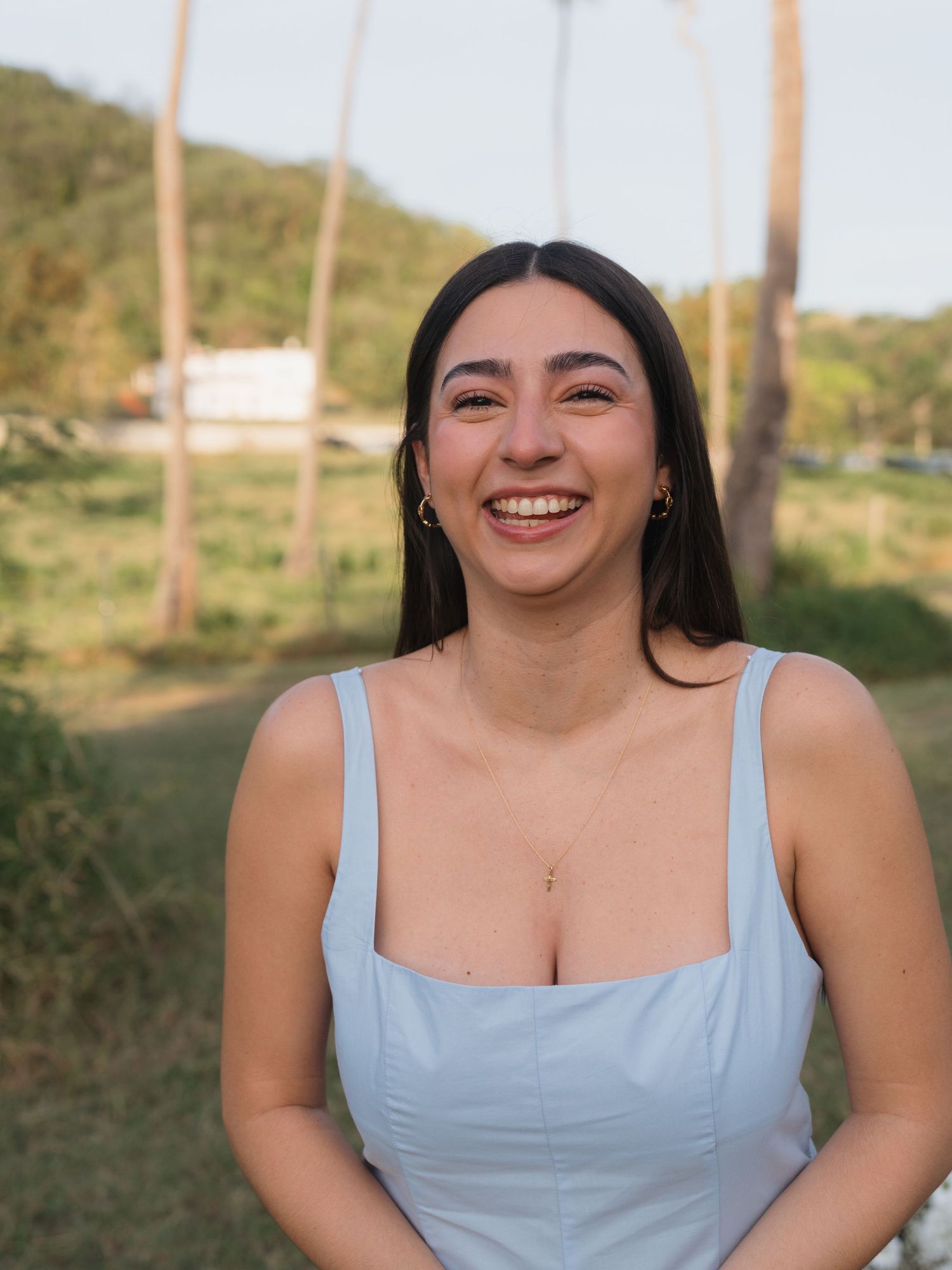 Smiling young woman in a light blue dress outdoors with green hills and palm trees in the background.