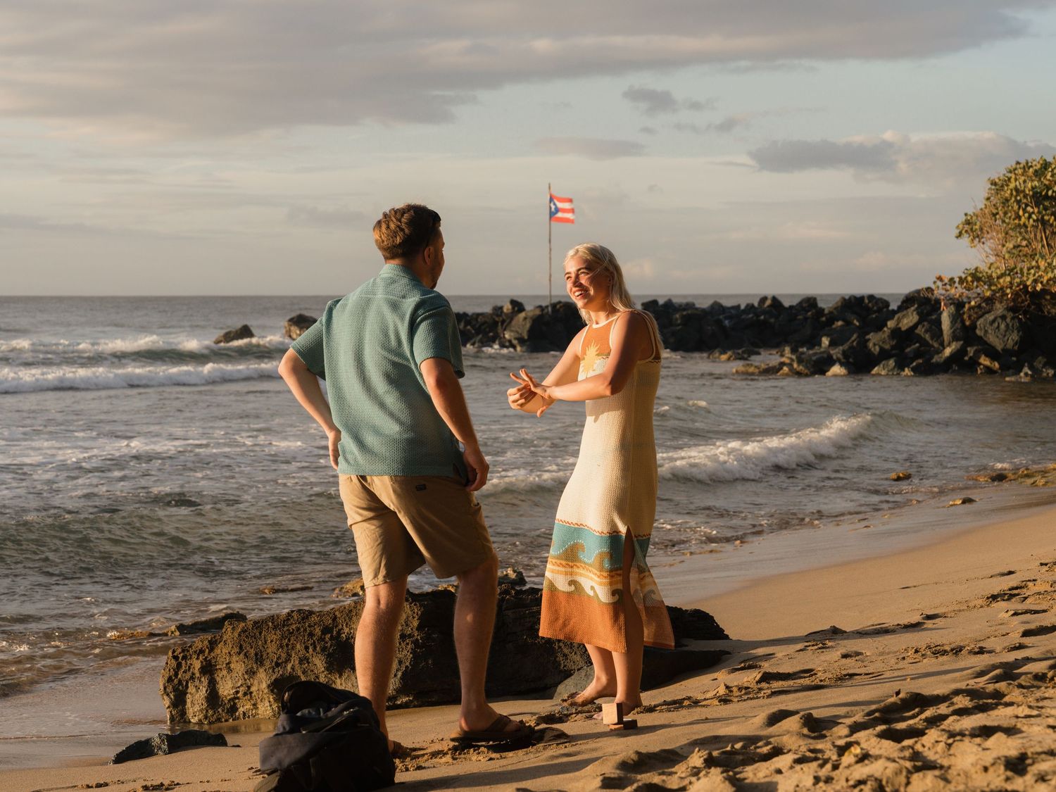 Couple standing on a tropical beach at sunset, with ocean waves and a Hawaiian flag in the background.