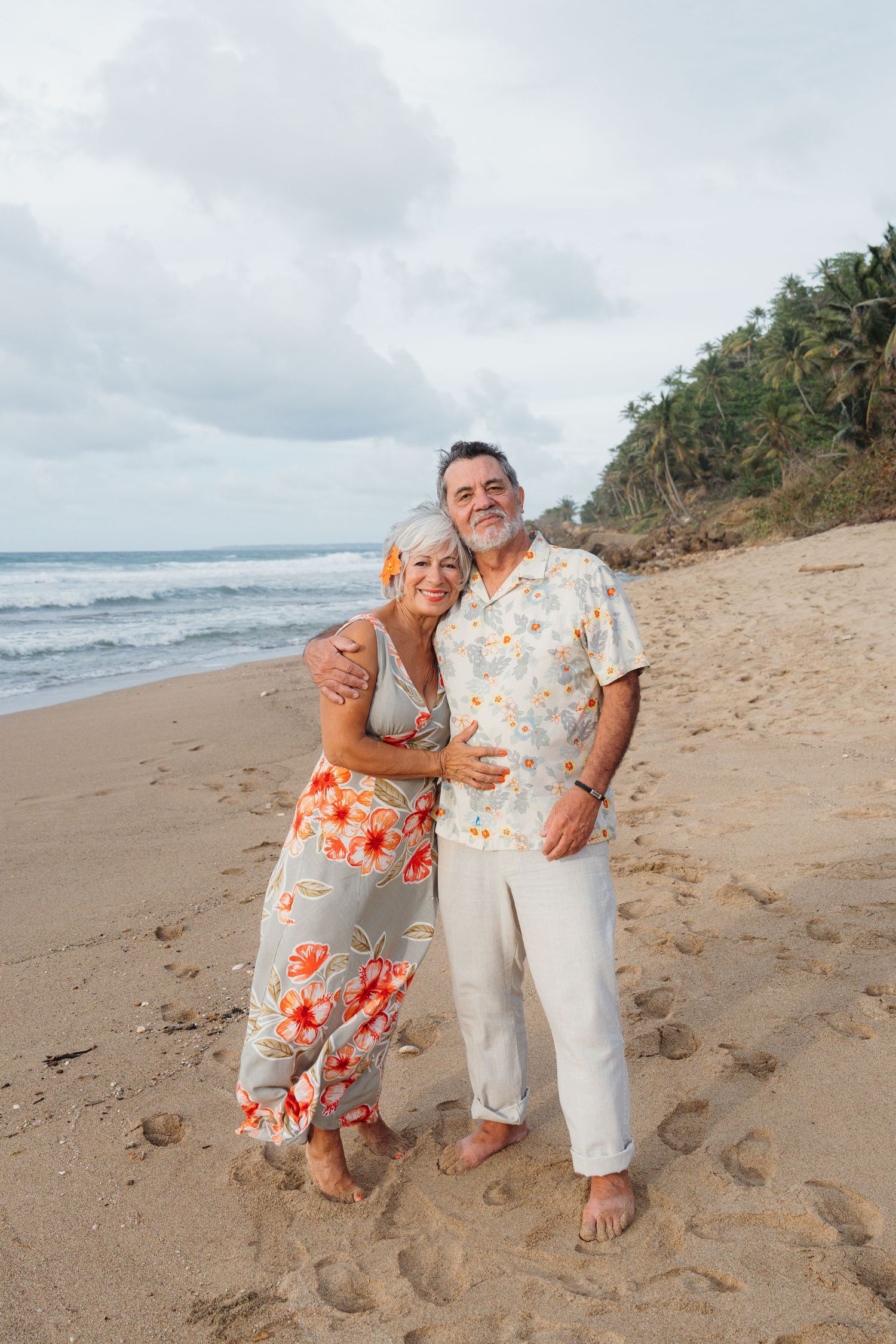 A happy senior couple embraces on a tropical beach, wearing floral outfits, with ocean waves and palm trees in the background.