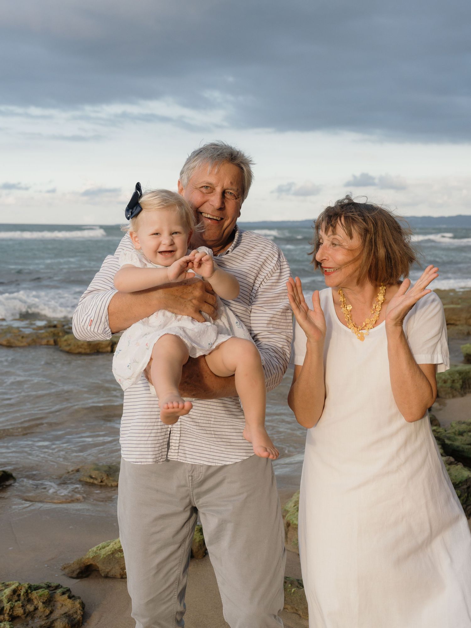 Grandparents laughing and playing with a baby girl on a rocky beach with ocean waves in the background.