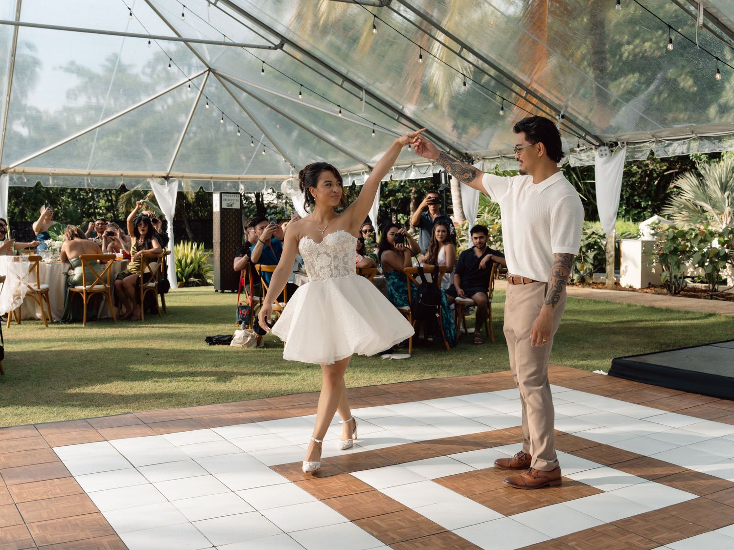 Couple dancing elegantly on outdoor wedding dance floor under a tent, woman in white tutu dress, man in casual attire.