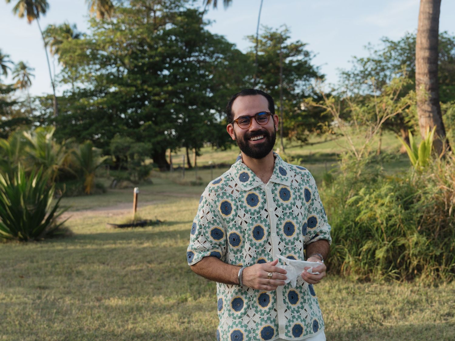 Smiling bearded man in floral shirt holding a cup outdoors in a tropical green landscape.