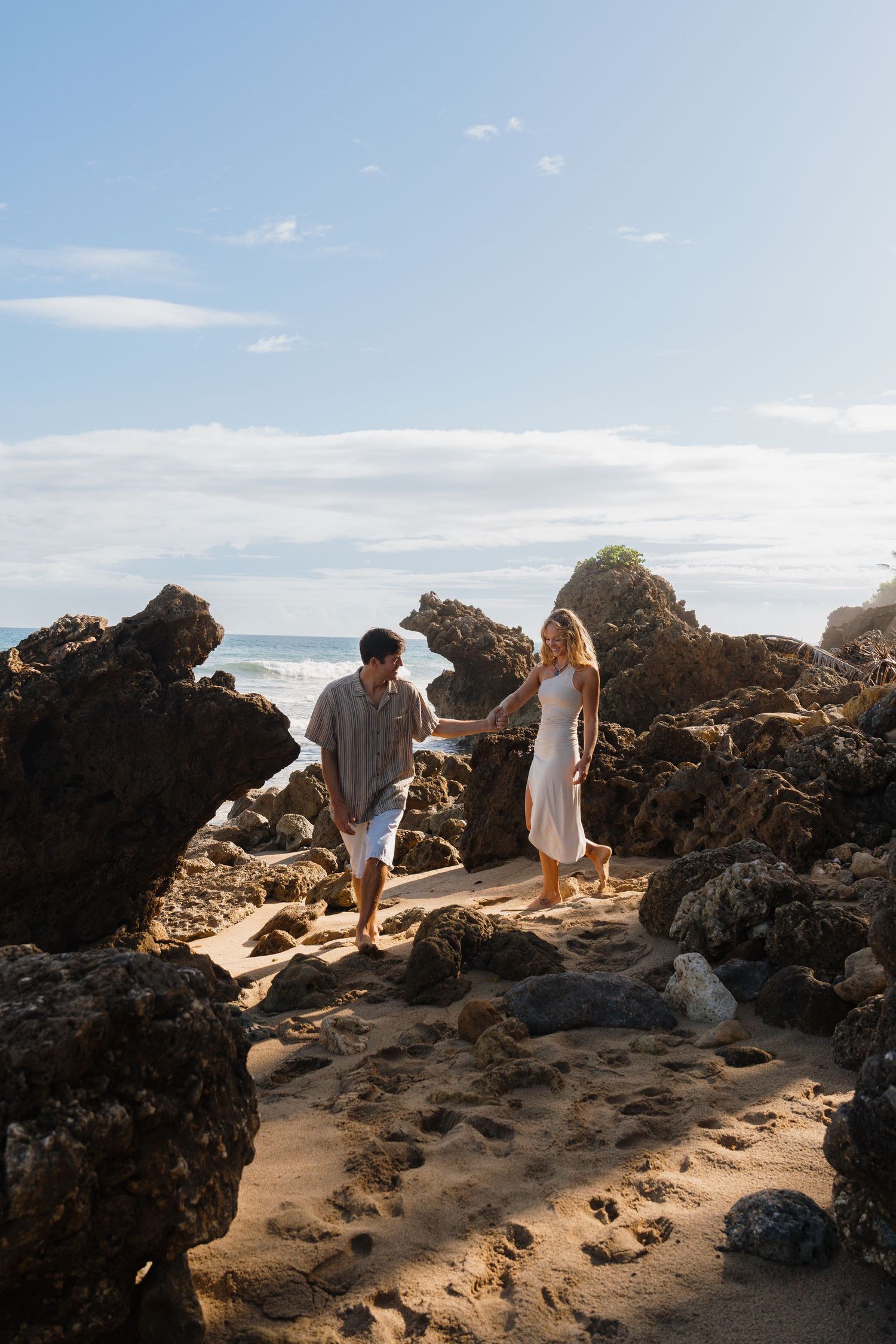 A couple holds hands while walking among rocky formations on a sandy beach with ocean waves in the background.