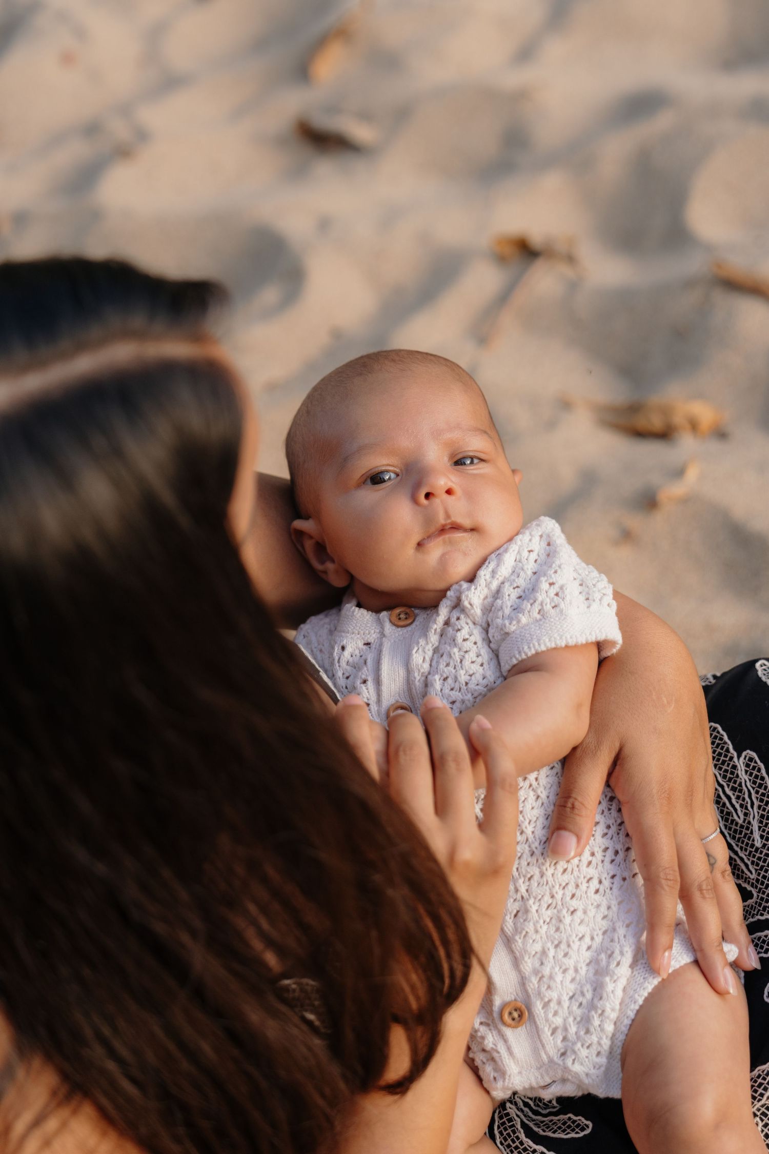 A mother holds her newborn baby dressed in white on a sandy beach in warm sunlight.