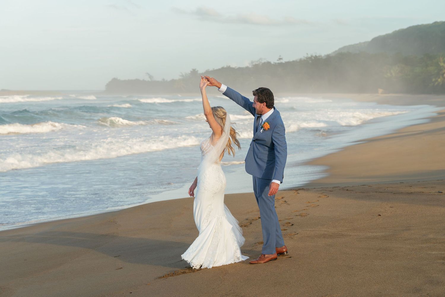 Bride and groom dancing on a tropical beach at sunset, waves crashing behind them.