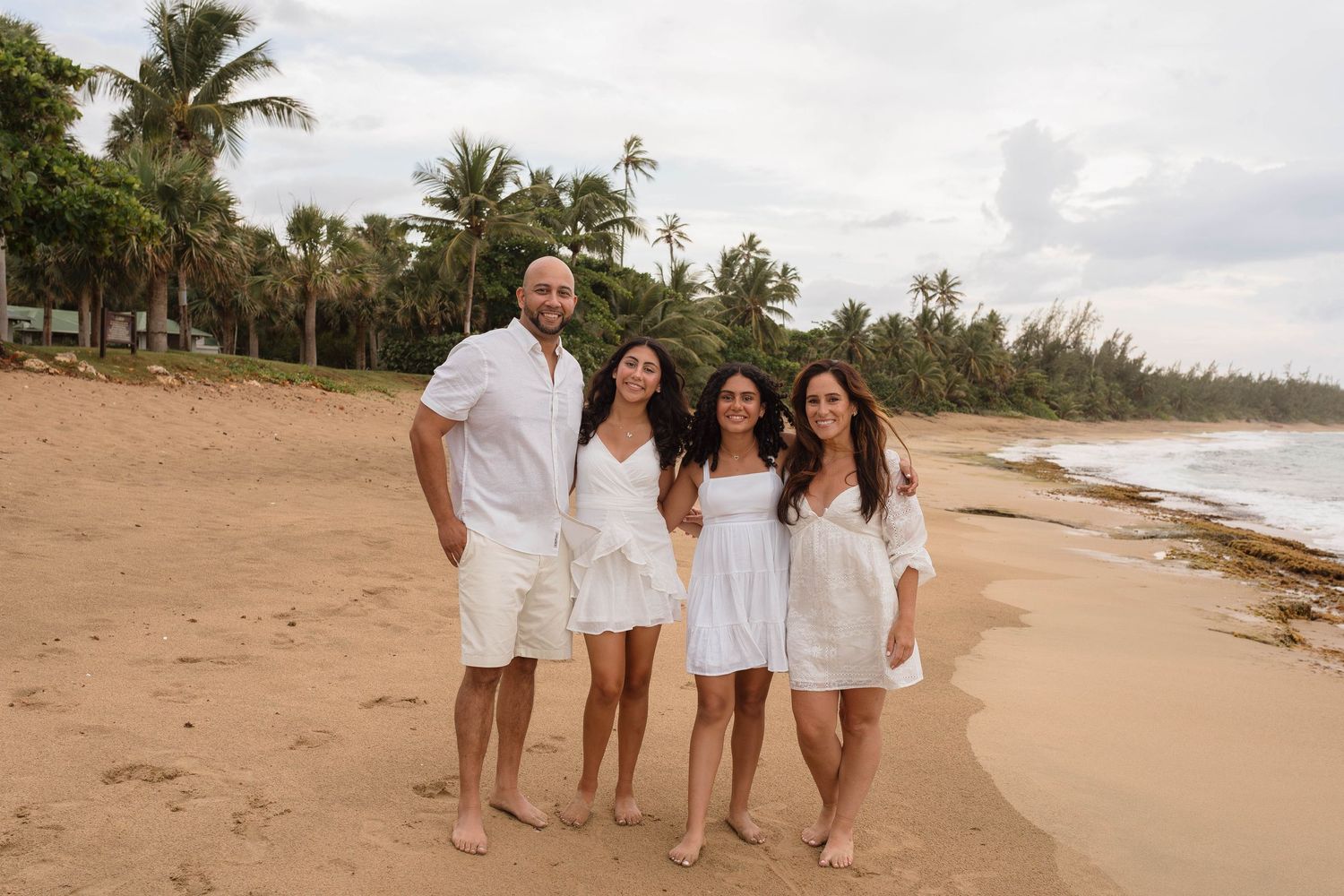 Family of four in white outfits smiling together on a tropical sandy beach with palm trees.