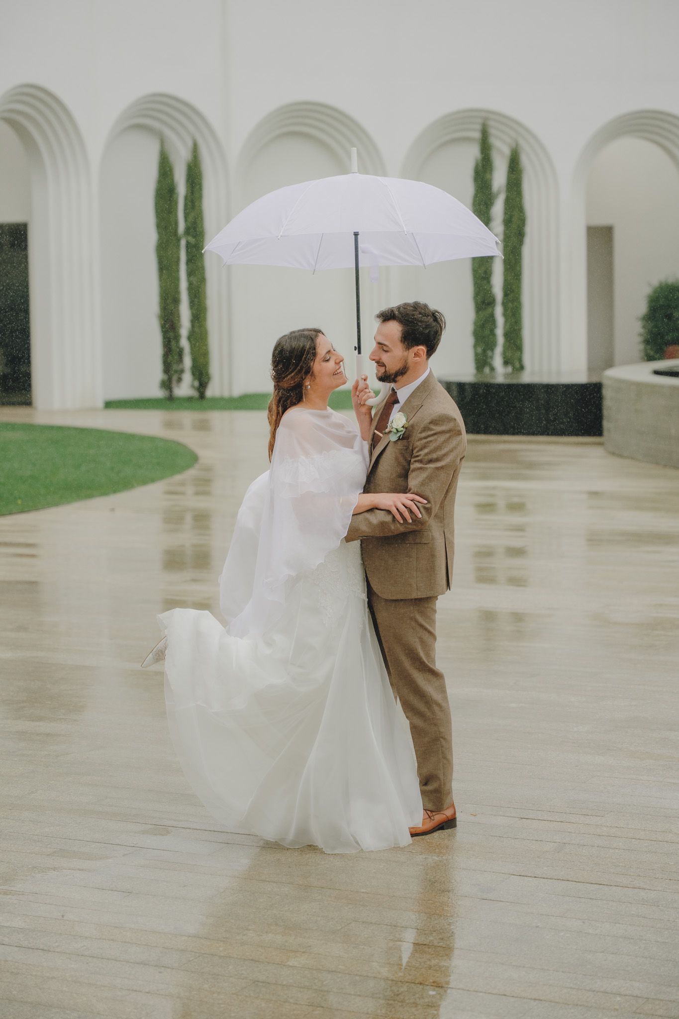 Couple sharing romantic moment with white umbrella in front of arched colonnade.