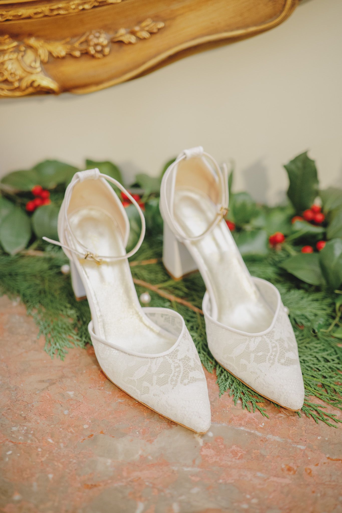 Elegant white bridal shoes displayed with holiday greenery and red berries.