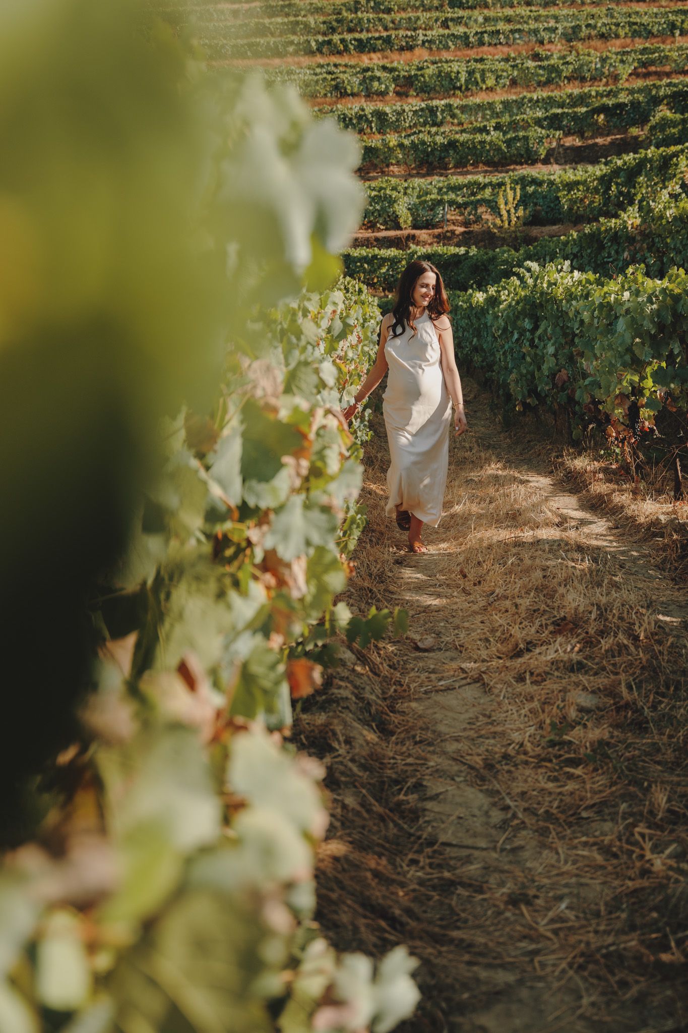 Woman in white dress walking through scenic vineyard rows during golden hour.