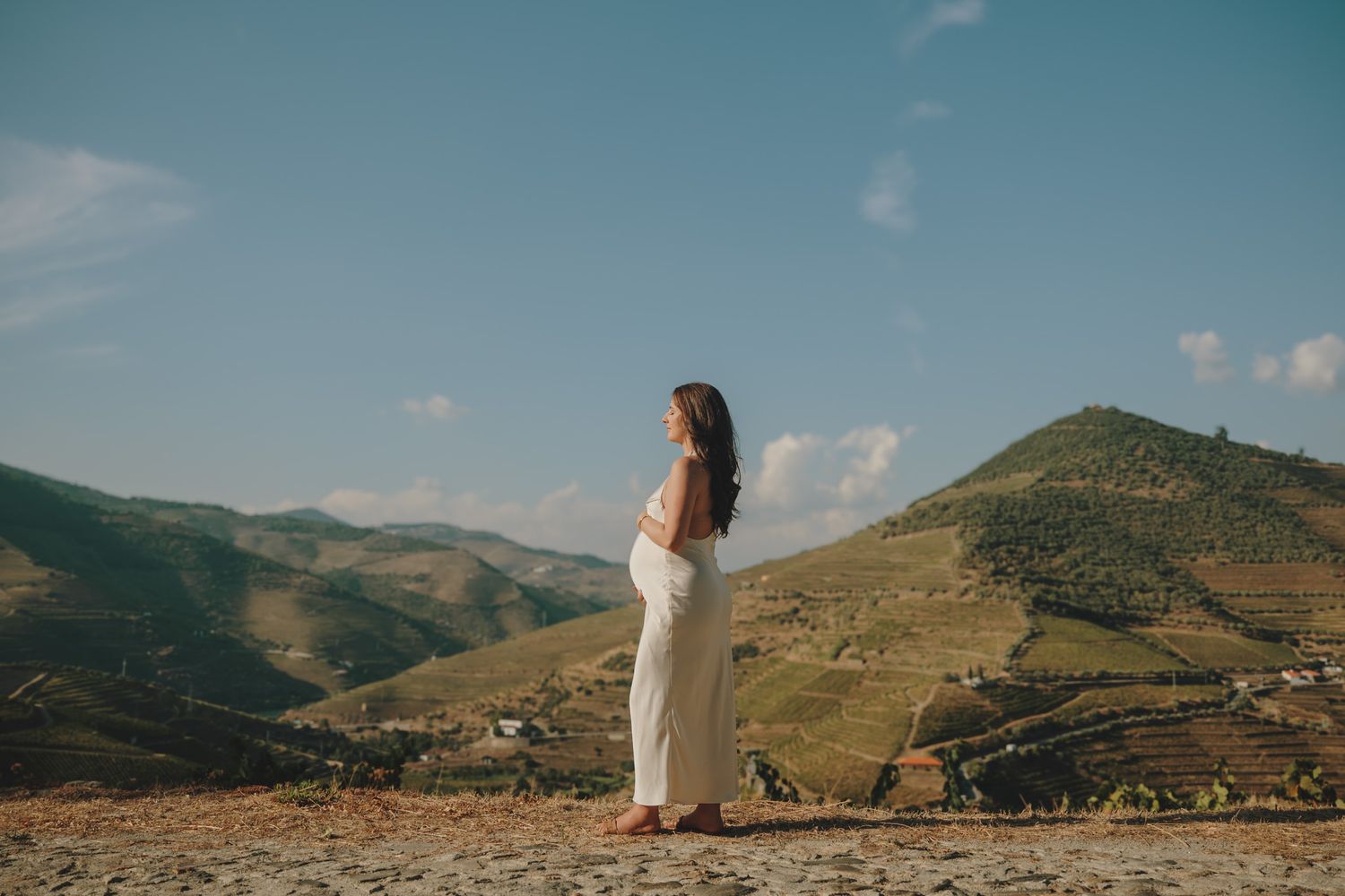 A figure in a white dress stands on a hilltop overlooking terraced vineyards in a scenic mountain landscape.