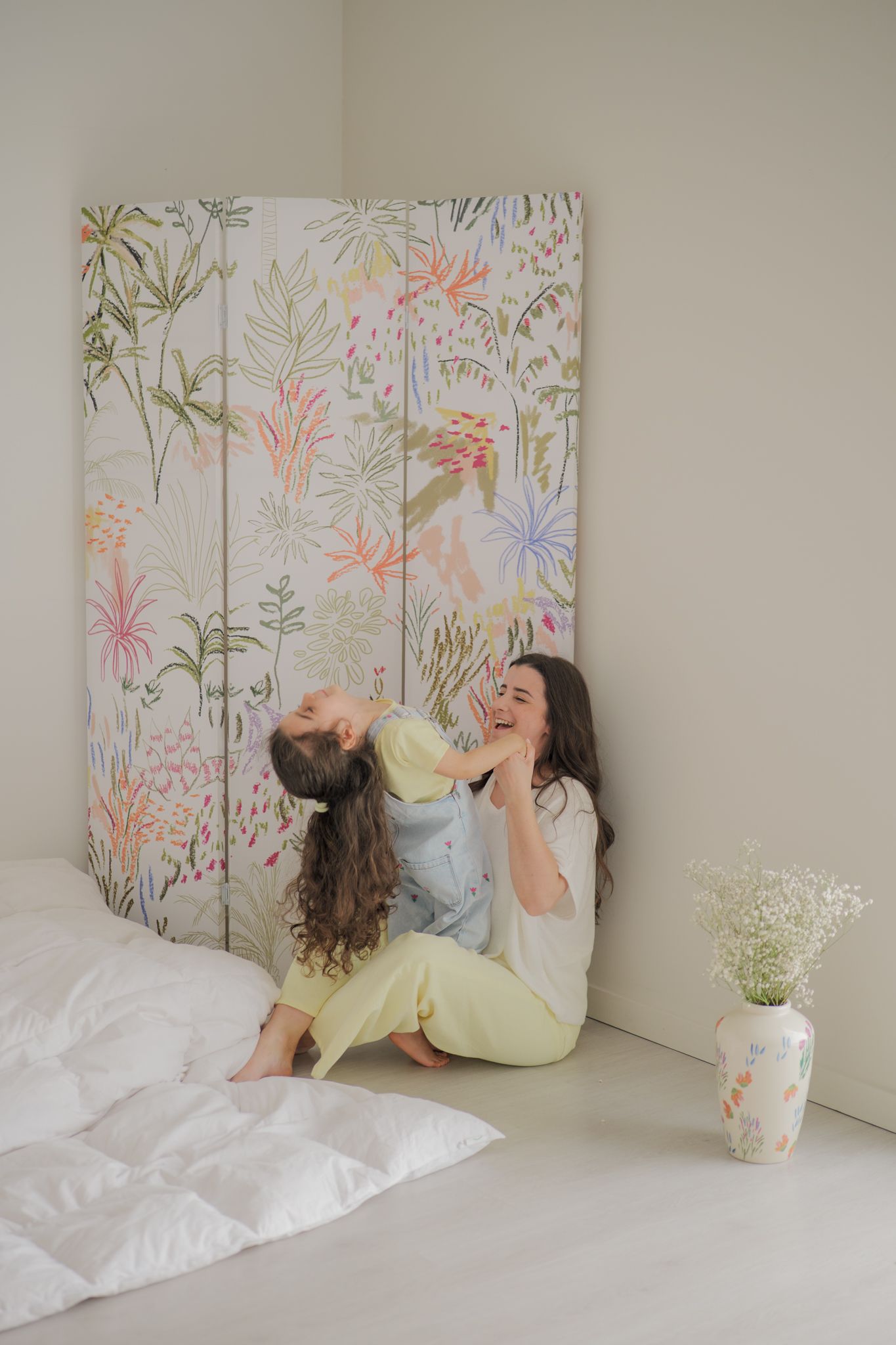 Mother and daughter laughing together on a white bed beside a floral decorative screen and vase.