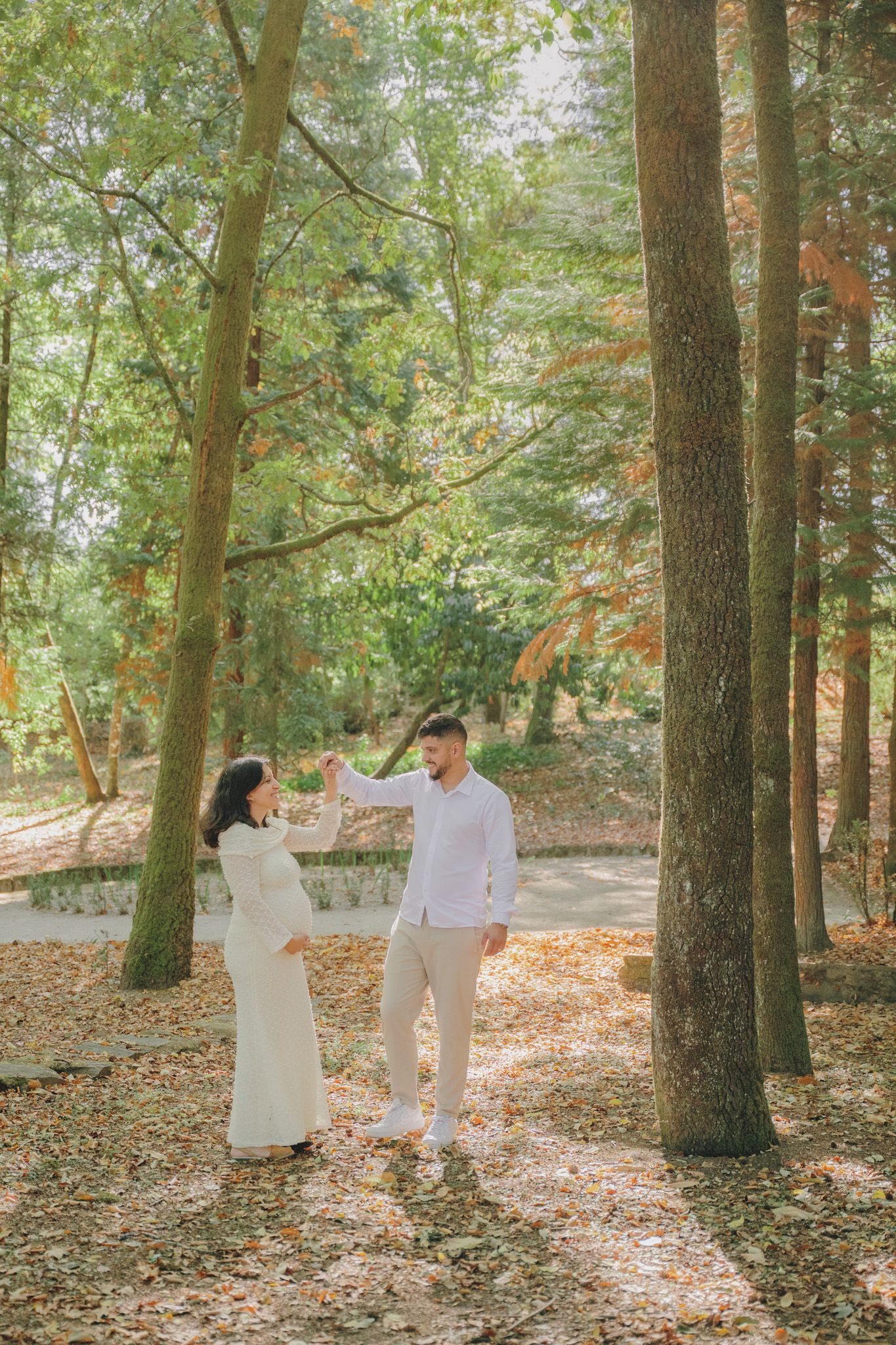 A couple in white outfits shares a tender moment on a wooded path covered in autumn leaves.