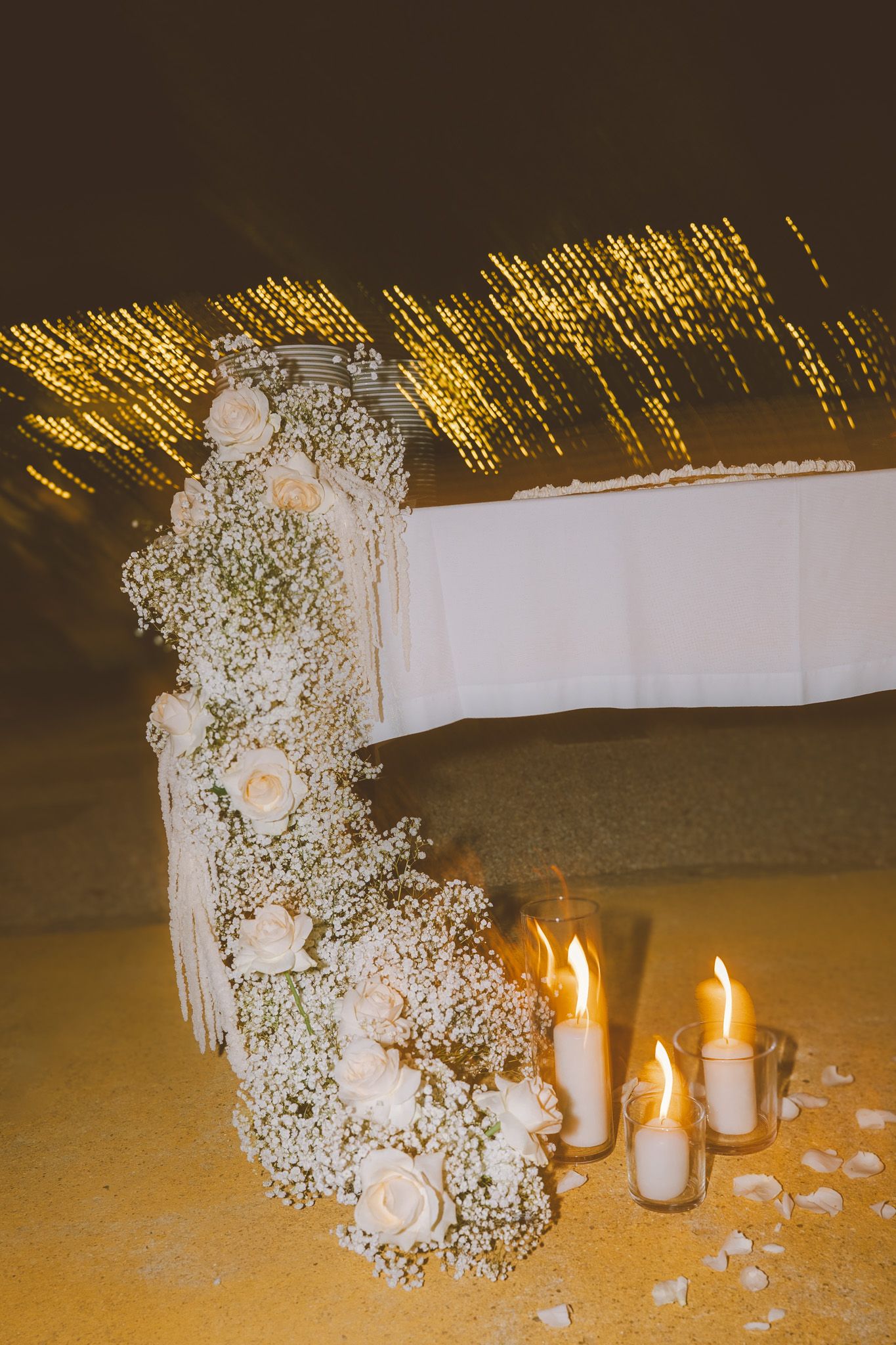 Elegant floral arrangement with baby's breath and roses decorated with glowing candles beneath string lights.