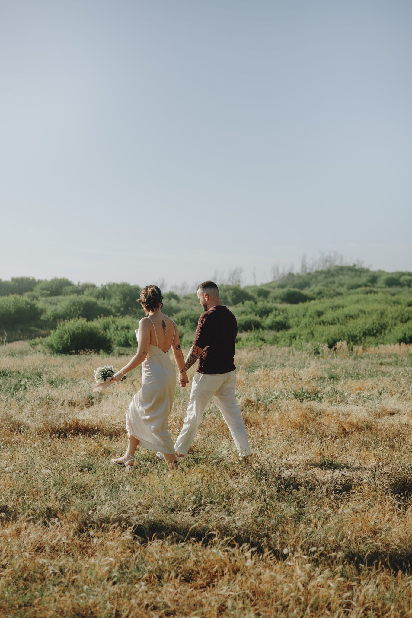Couple walking through a sunlit field holding hands in coordinated white outfits.