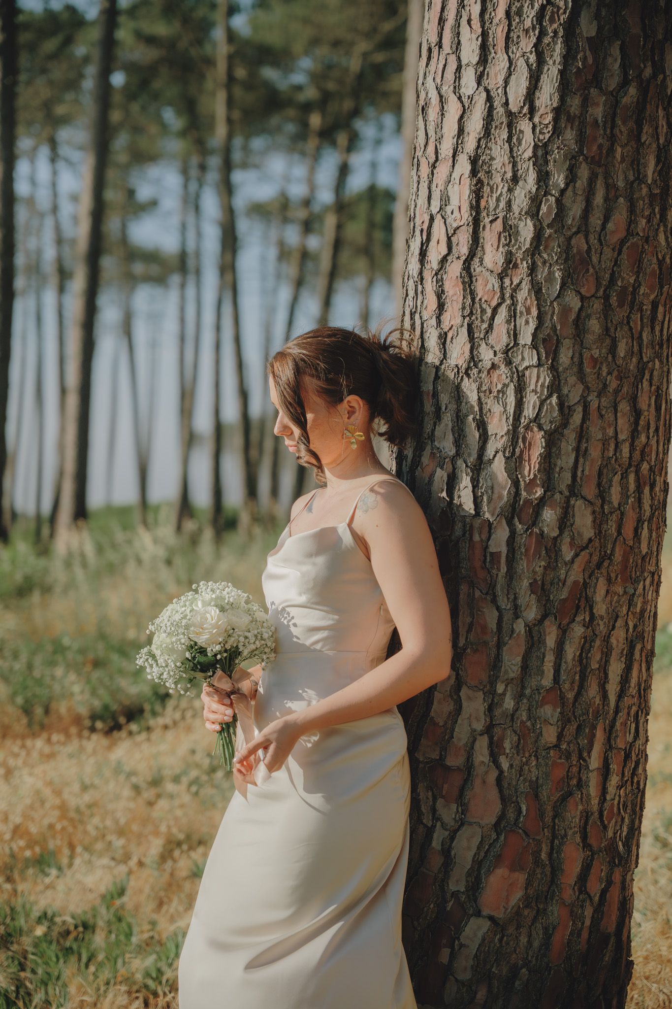 Bride holding white bouquet poses against pine tree trunk in forest setting.
