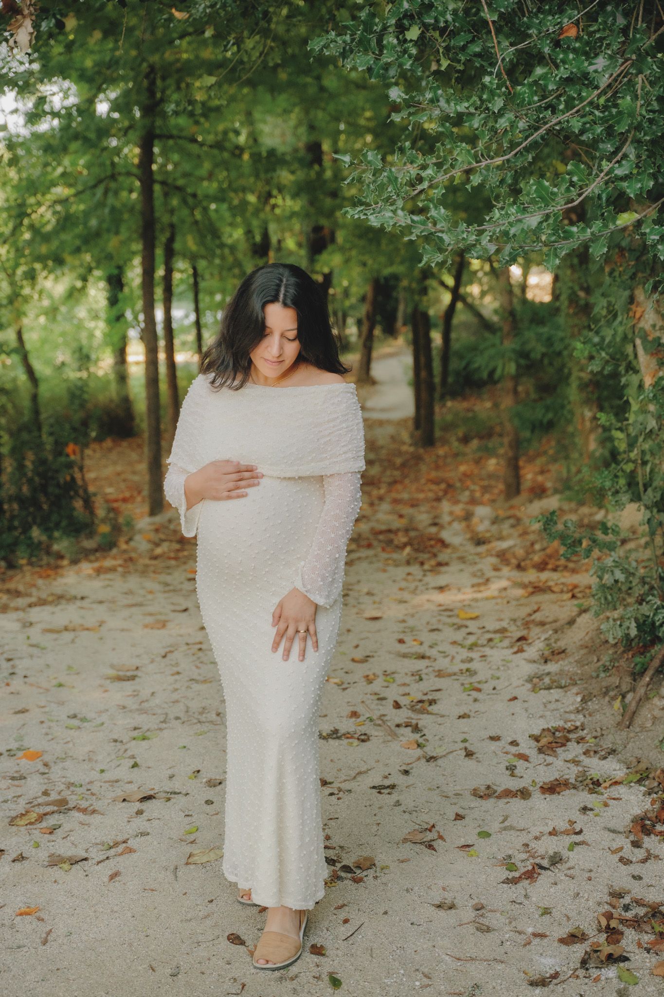 Maternity photo on a leaf-covered path featuring a white off-shoulder dress.