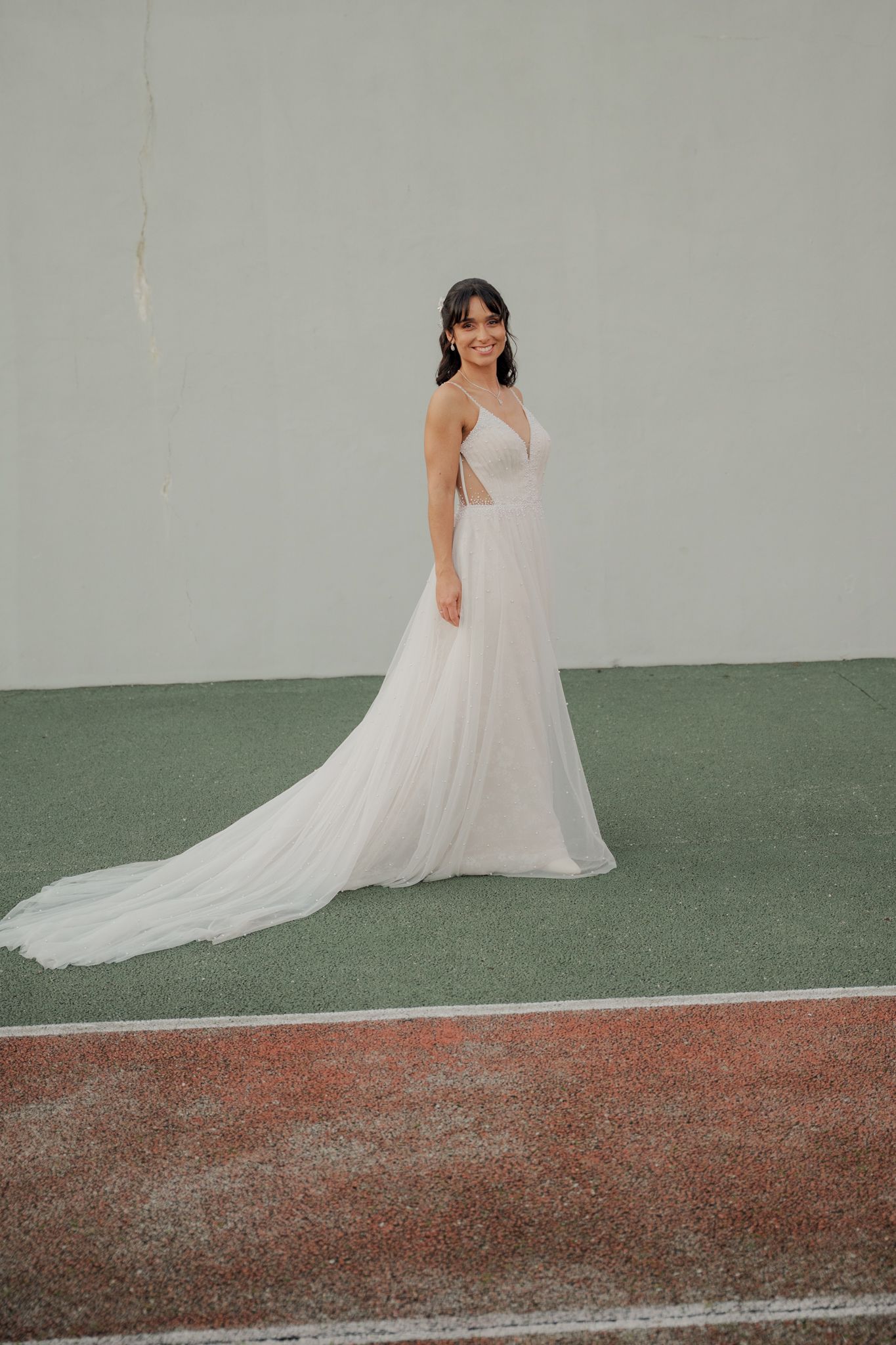 Bride in flowing white gown poses elegantly on sports court during wedding portraits.