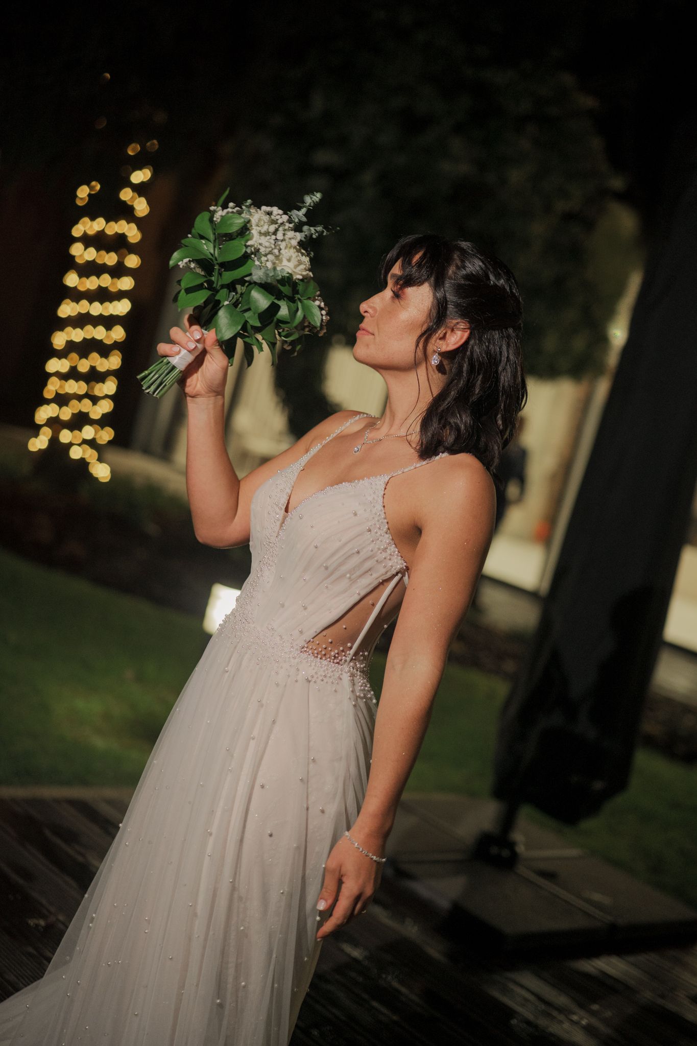 A bride in a flowing white dress enjoys a drink at an outdoor evening reception.