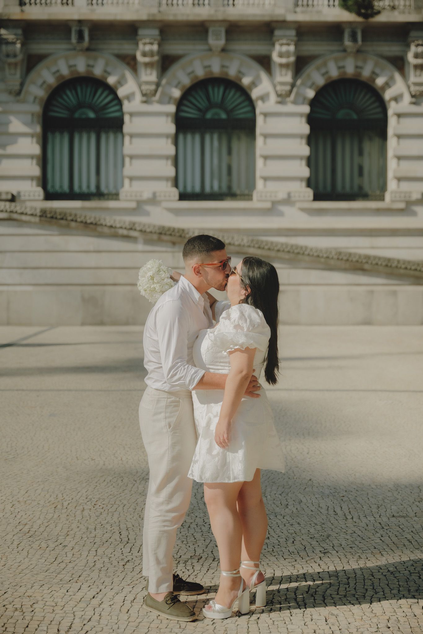 Romantic couple in white attire embracing on cobblestone plaza near historic building.