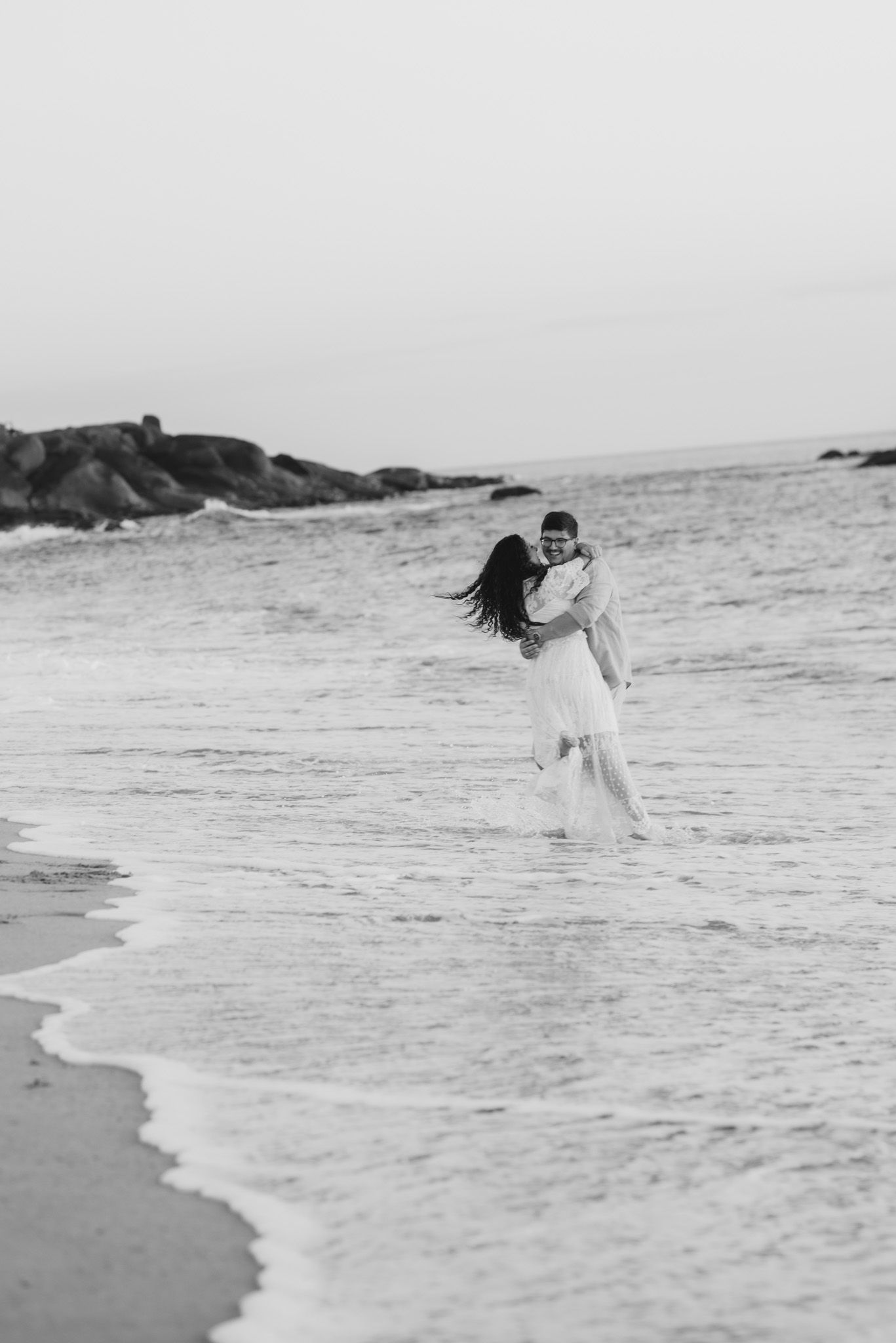 A couple splashes in the ocean waves wearing formal attire in black and white.