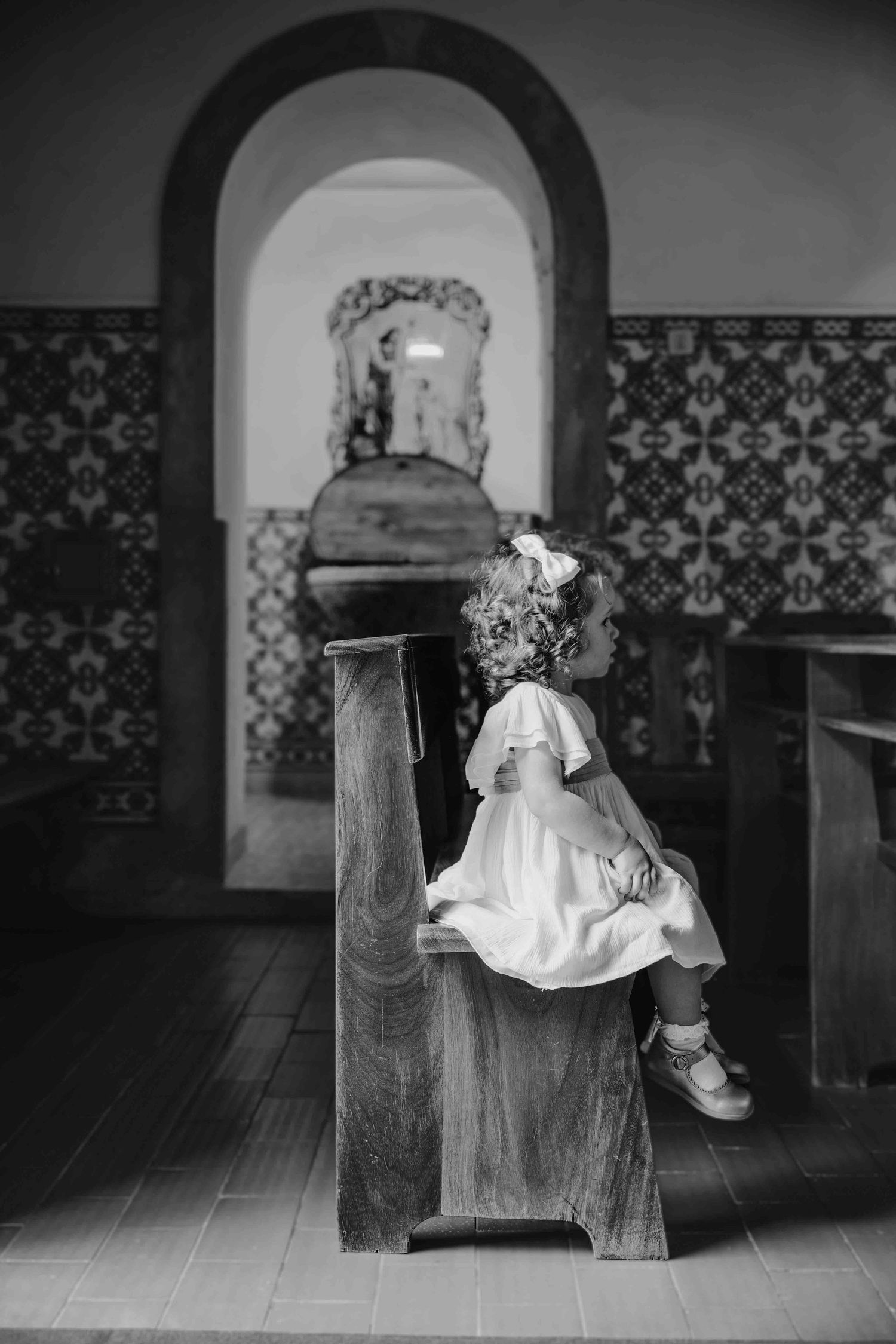 A child in a white dress sits on a wooden church pew, looking up at an arched doorway with decorative tiled walls.