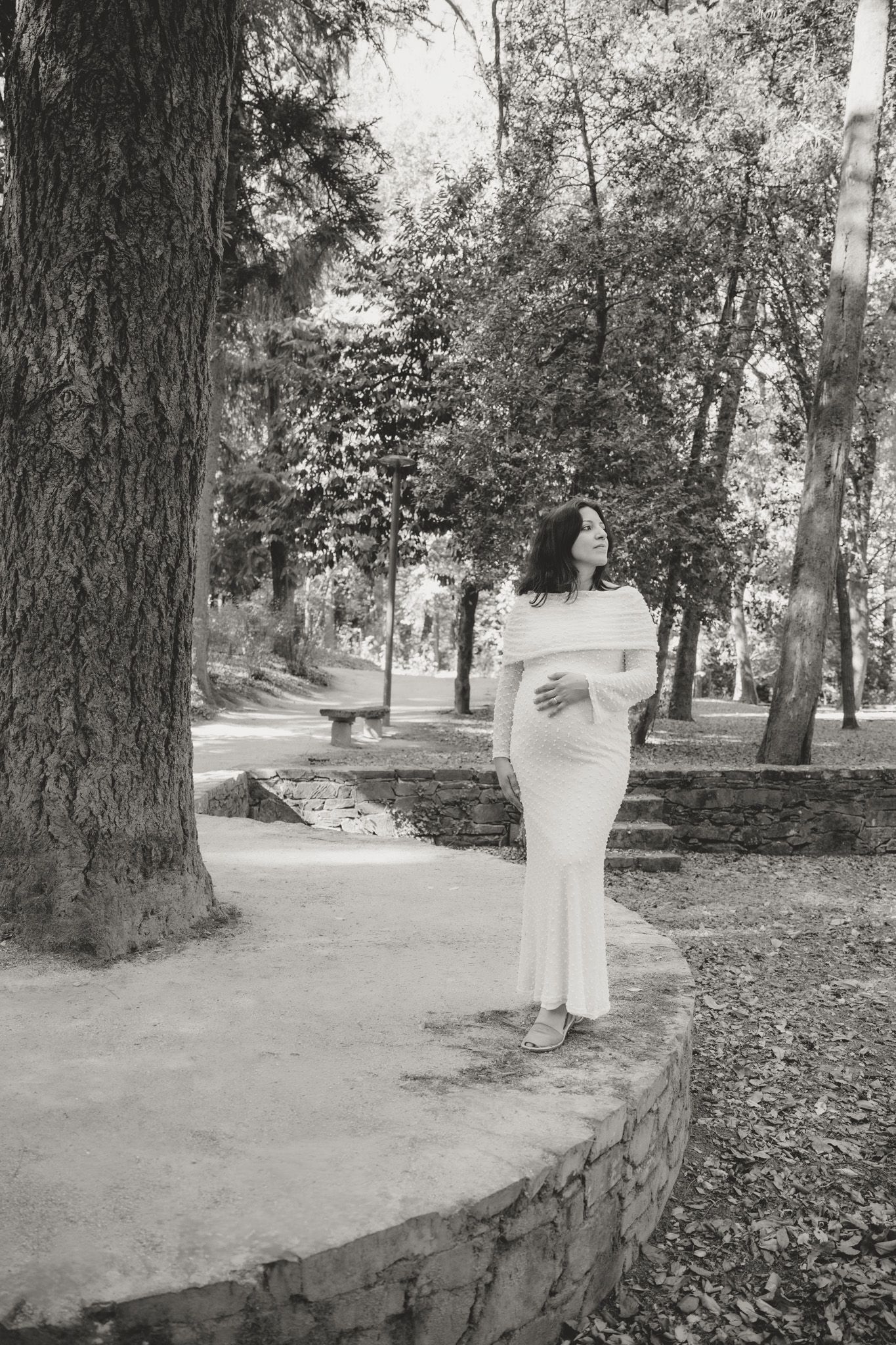 A woman in a flowing white dress poses on a stone path surrounded by trees.