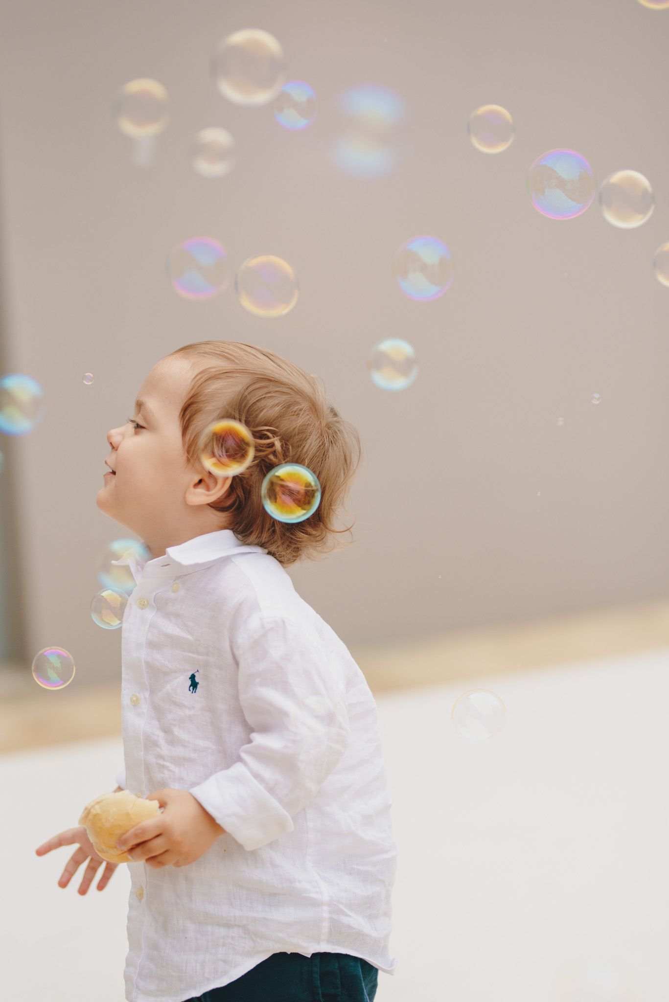 Child in white clothing chasing and playing with floating soap bubbles.