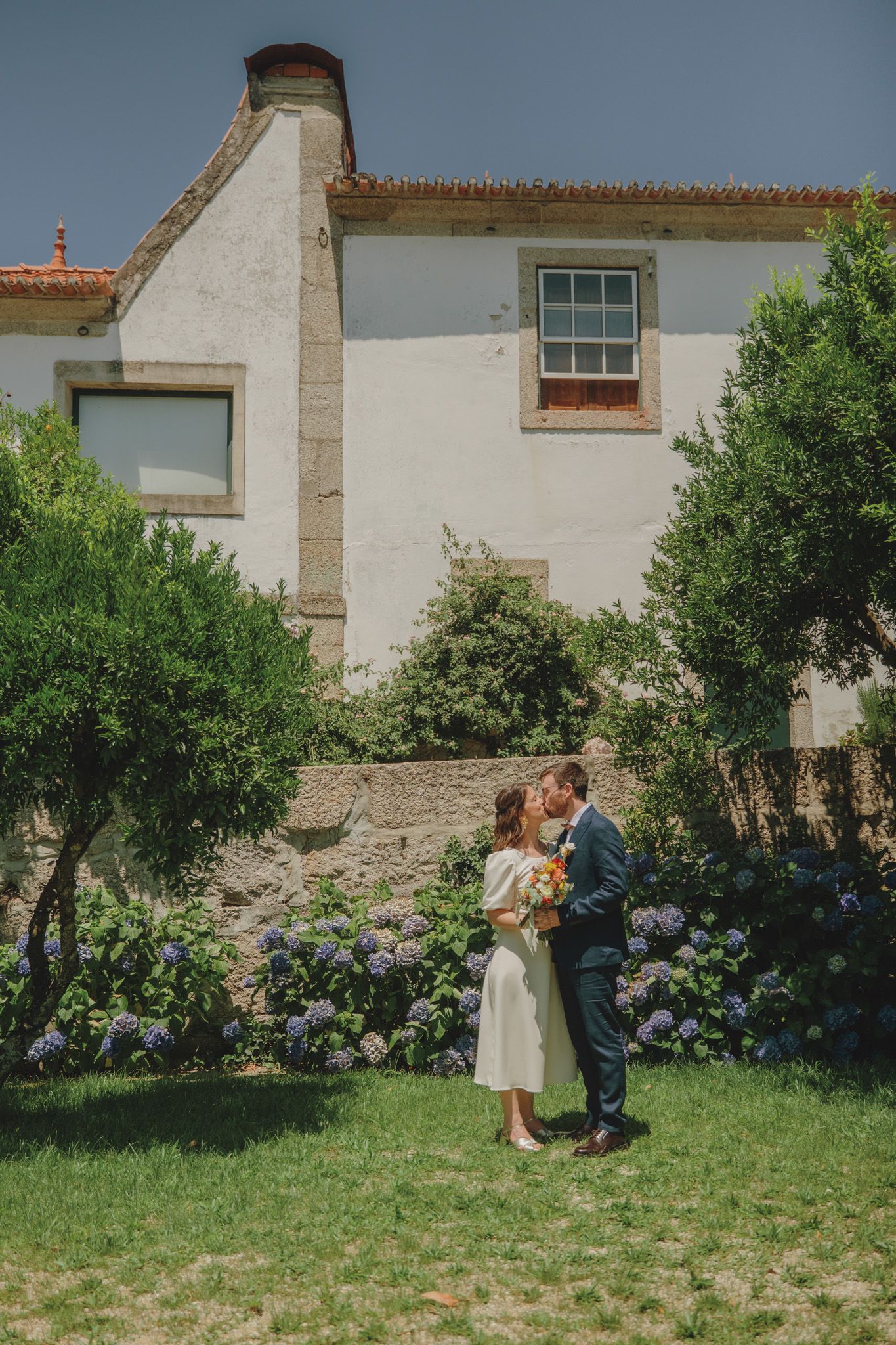 Wedding couple sharing intimate moment in garden with white building backdrop.