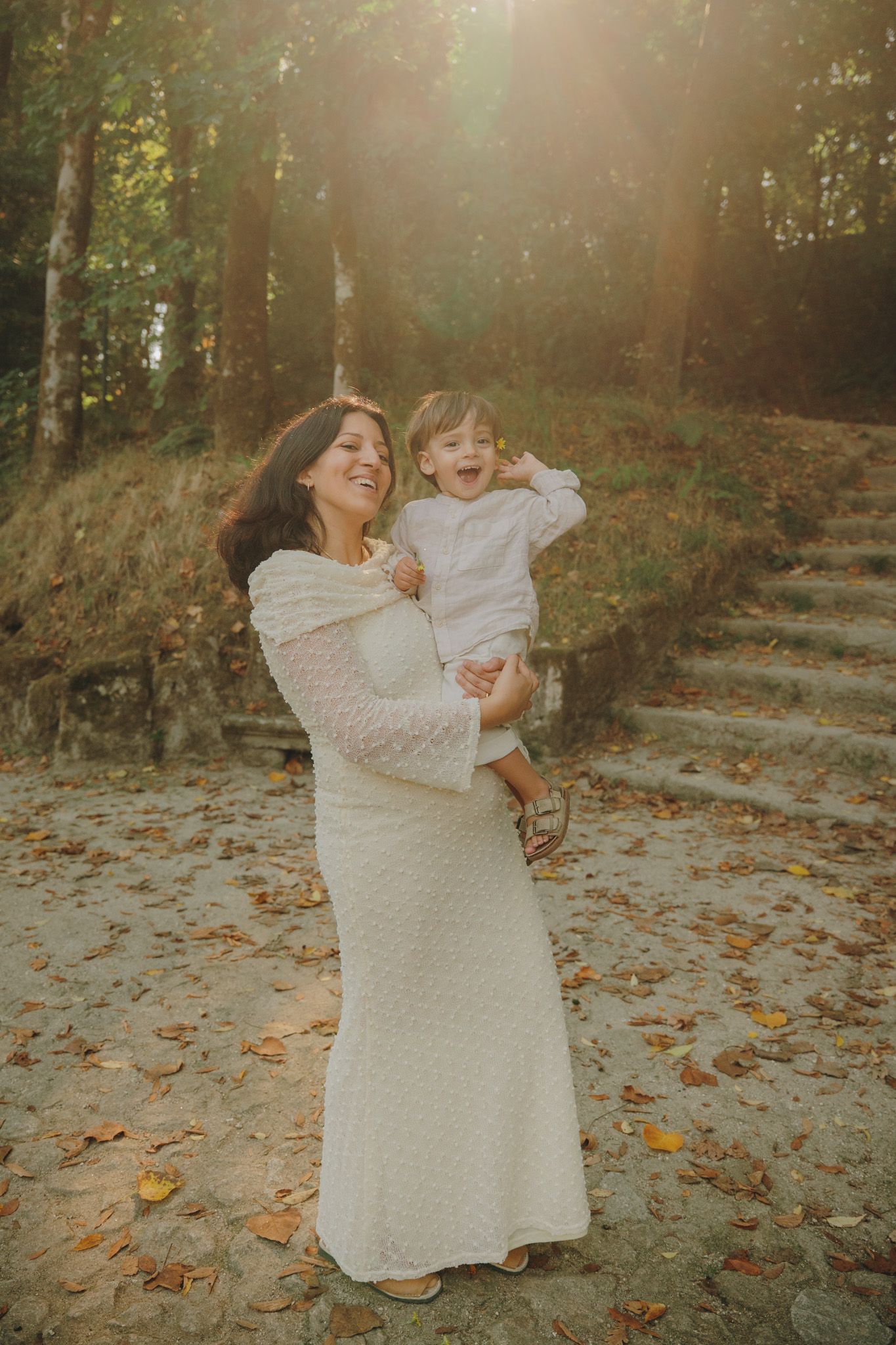 Mother and child in matching white dresses share joyful moments while playing in a sunlit autumn forest setting.