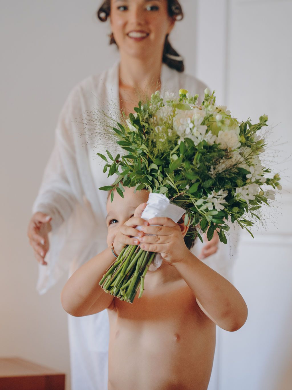 A person in a white top holds a fresh green and white floral bouquet in a bright indoor setting.