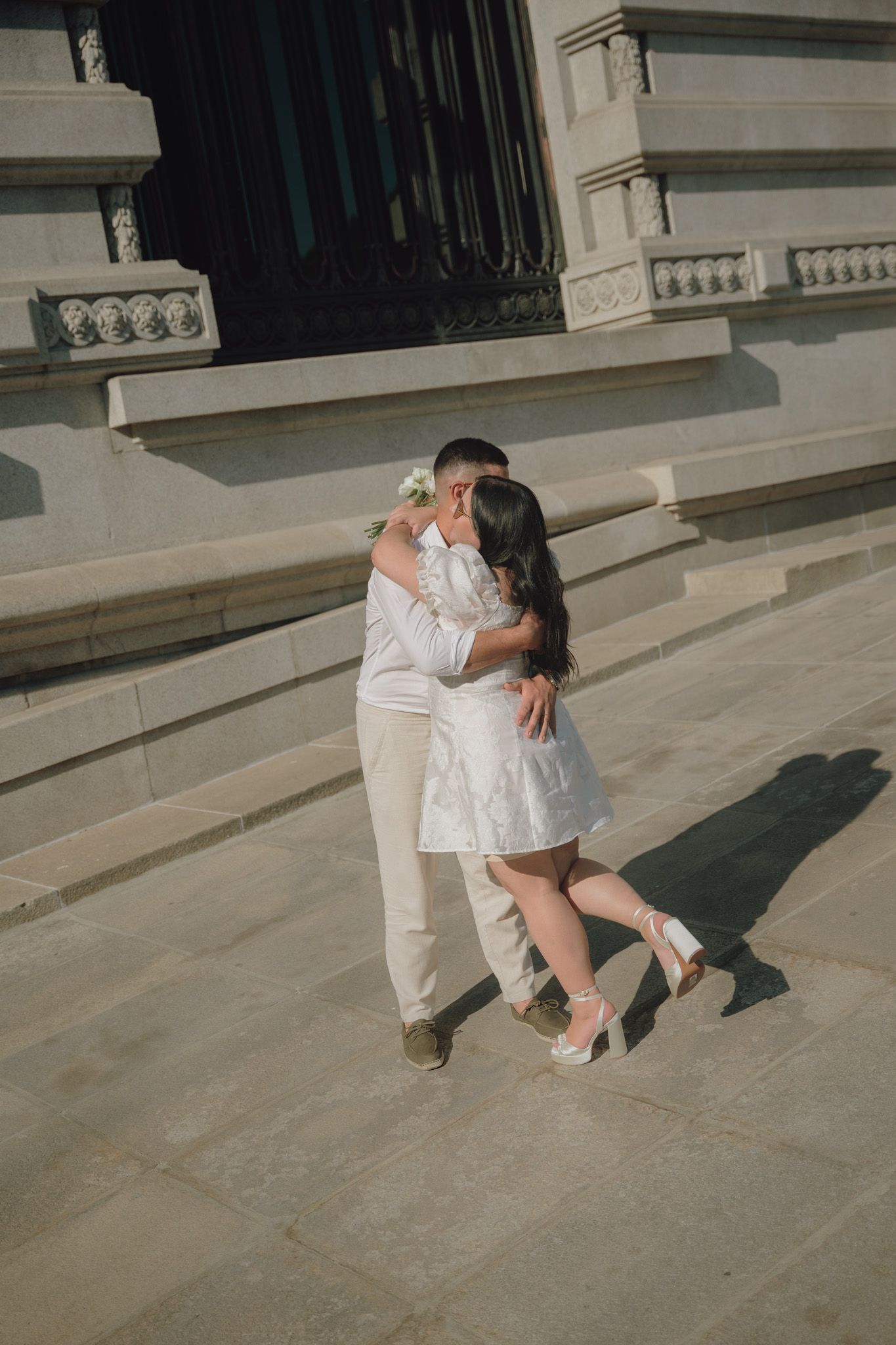 A couple in white outfits poses playfully on stone steps in sunlight.